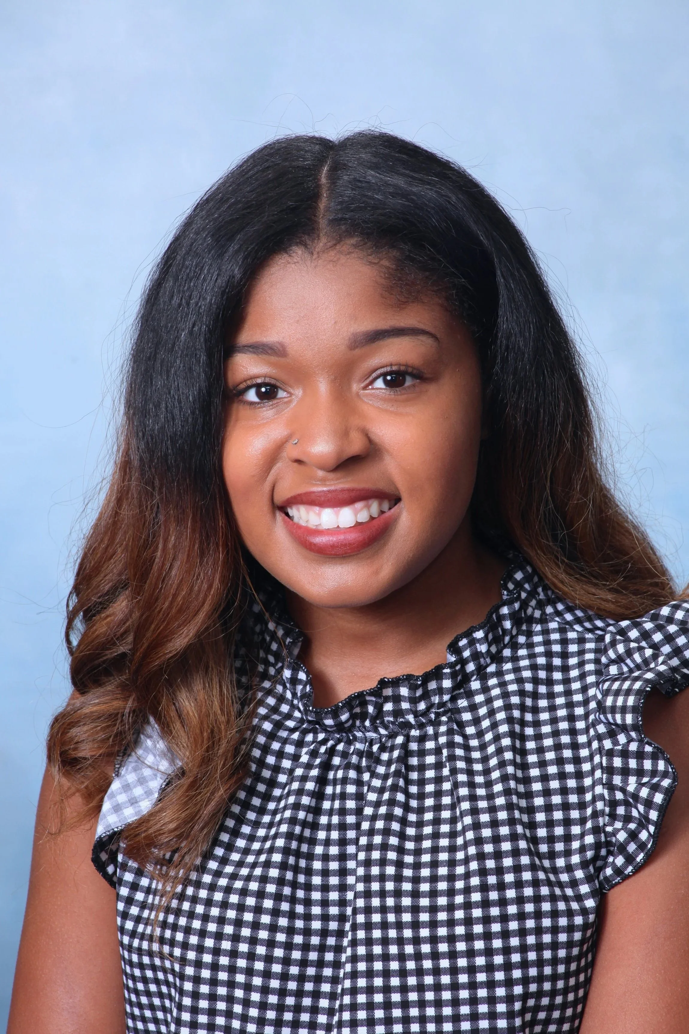 Close-up portrait of a young African American woman with long, curly hair, smiling, wearing a black and white checkered dress, against a light blue background.