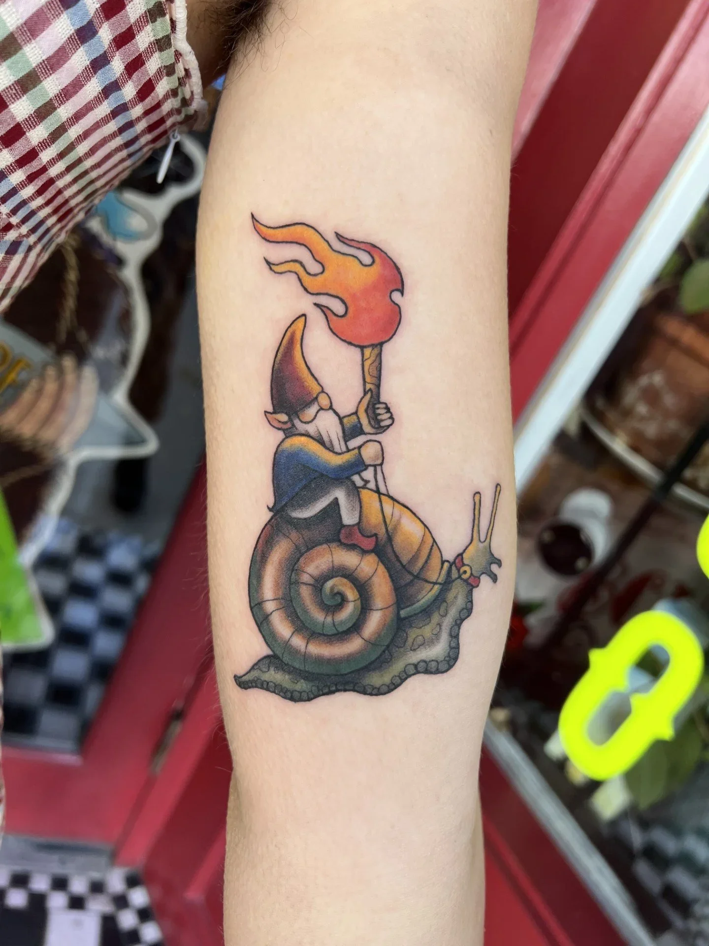 A colorful tattoo on a person's arm features a snail riding a bike with a flame-headed character holding a torch, set against a checkered background.