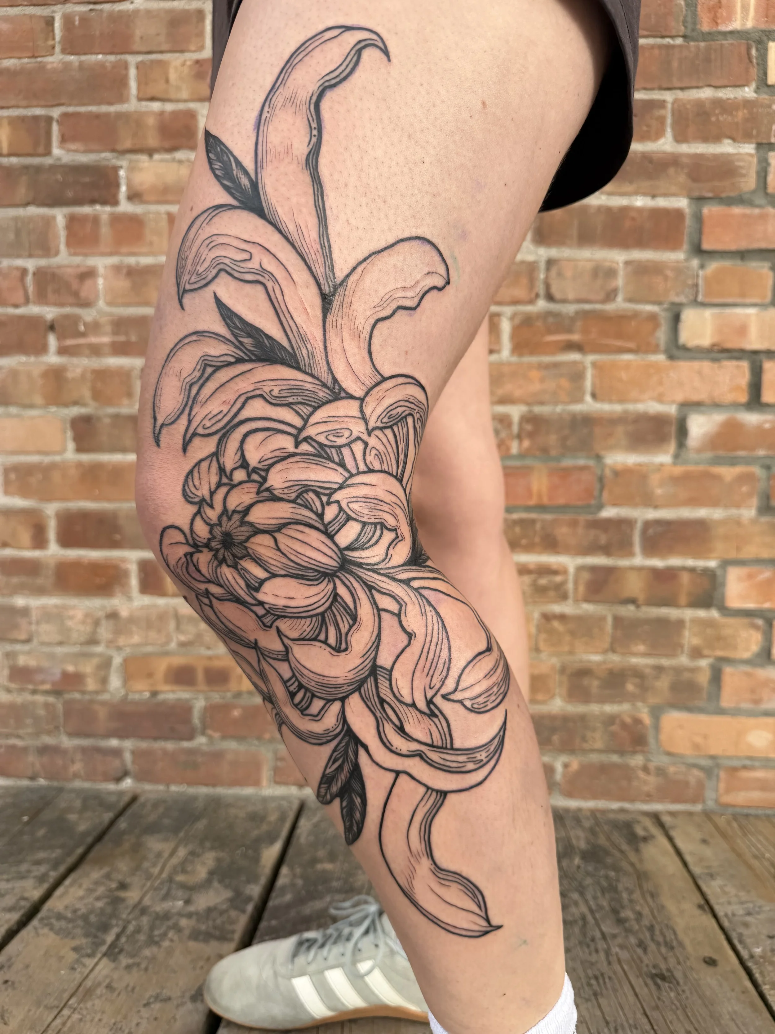 A tattoo of a flower with large petals and long leaves, extending from the upper thigh to the lower leg, with detailed line work, on a person's smooth skin. The background features a brick wall and wooden floor.