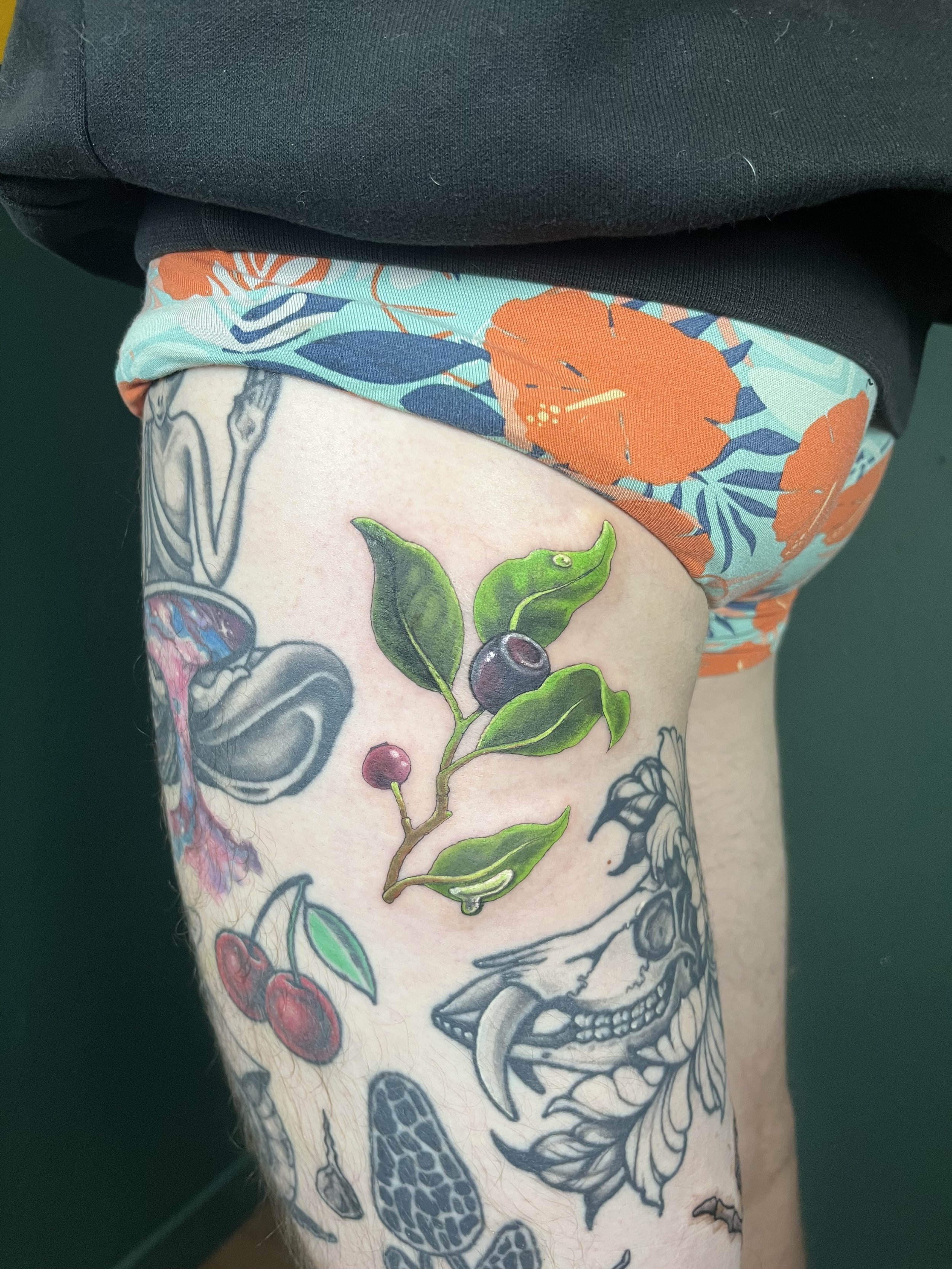 A tattoo of a skull with a detailed design on a person's arm, surrounded by colorful floral and botanical elements, including cherries, leaves, and flowers.