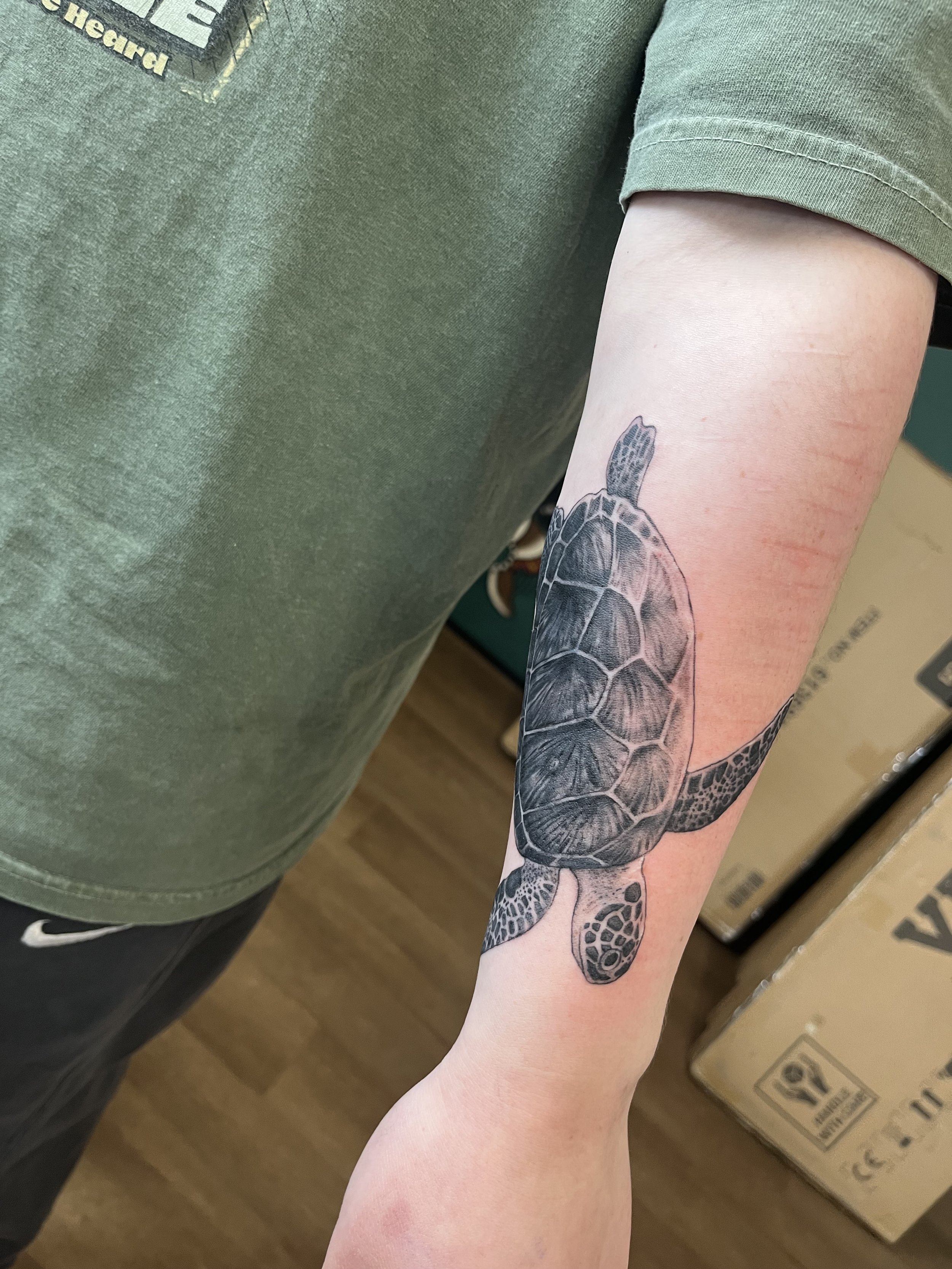 The image shows a person's arm with a detailed black and gray tattoo of a turtle, including the shell and limbs. The person is wearing a green T-shirt with a patch and is standing indoors.