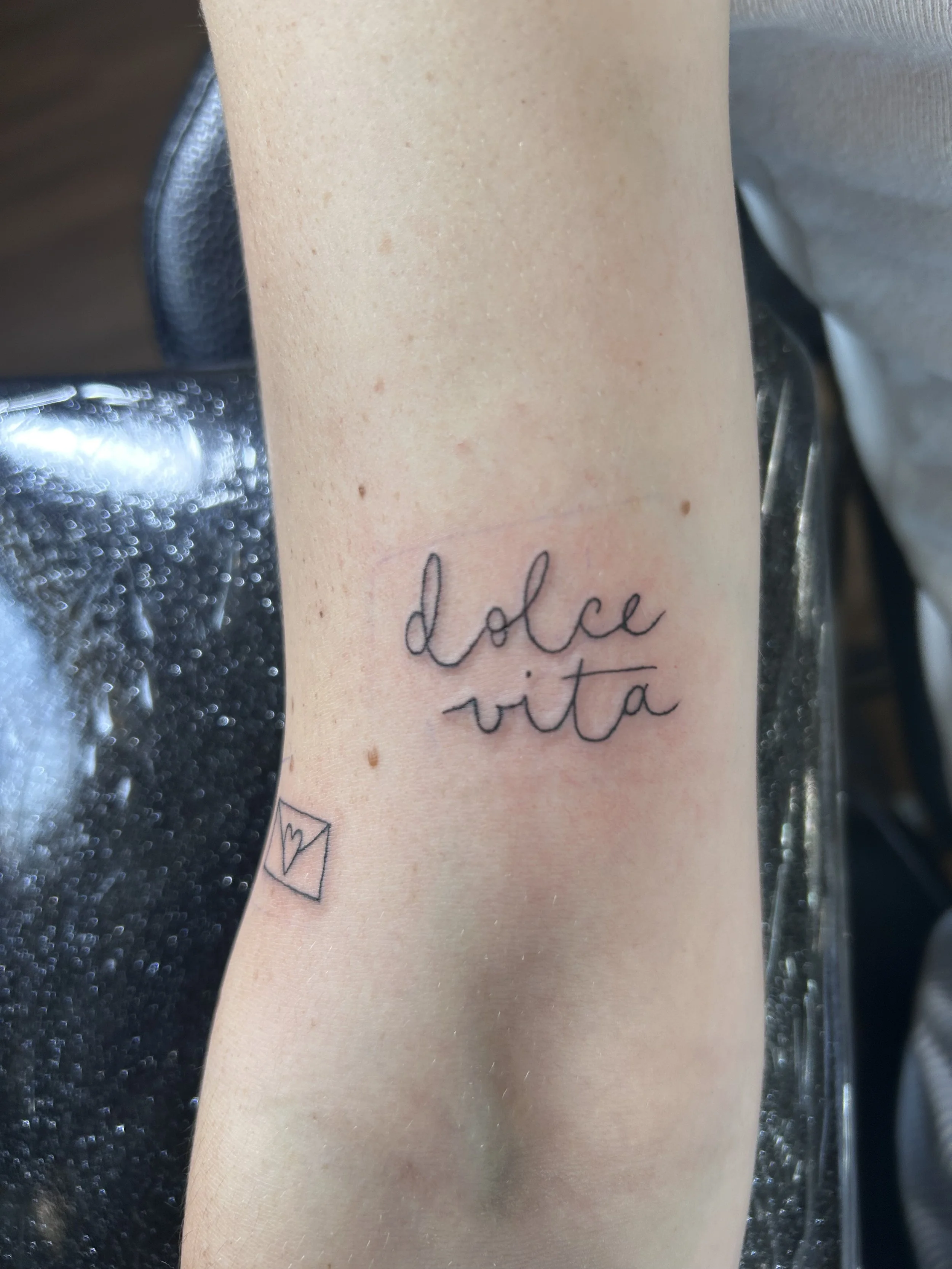Close-up of a person's forearm with a tattoo of the words 'dolce vita' in cursive and a small open envelope with a heart inside underneath the text.