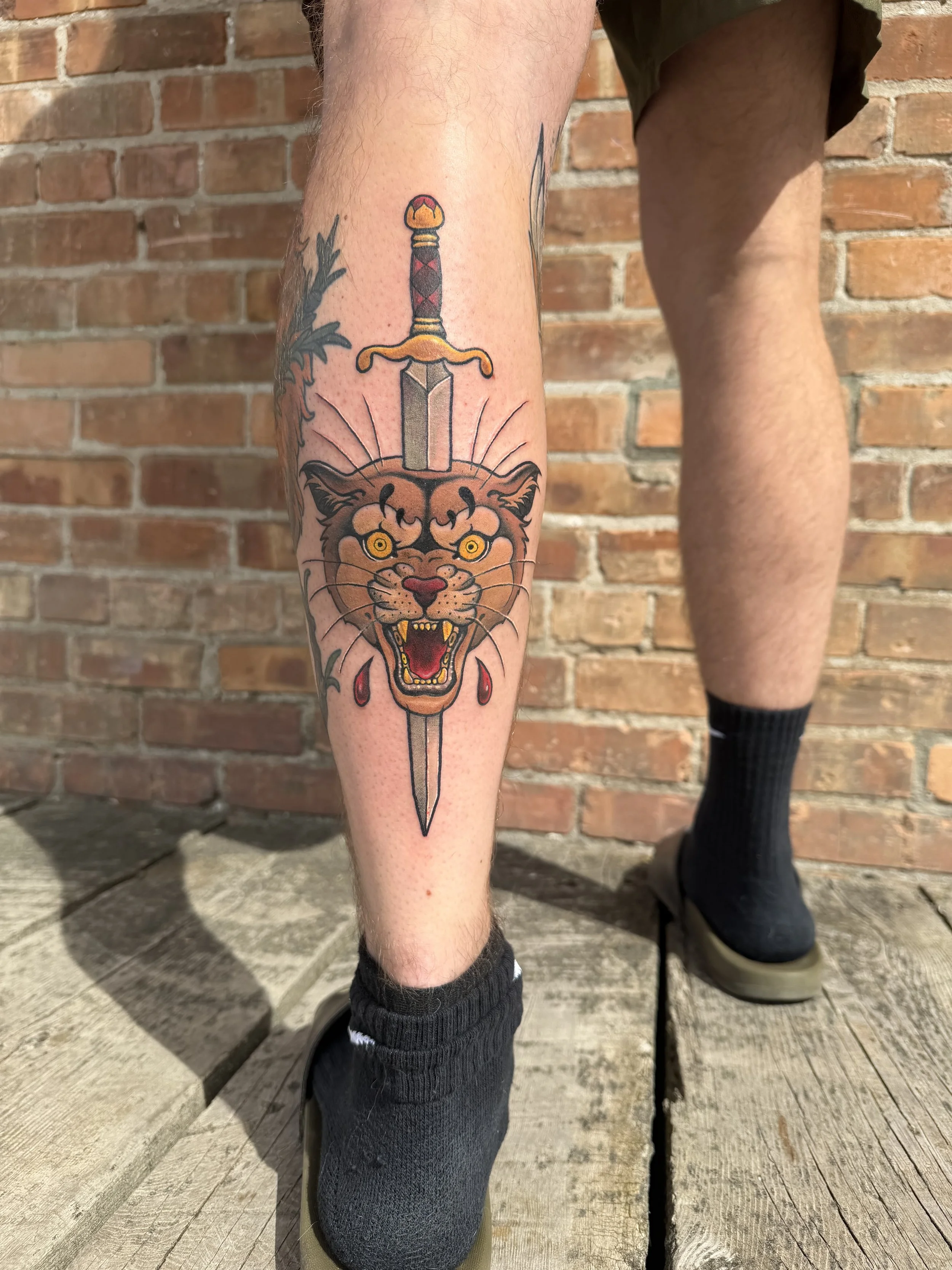 Tattoo of a fierce lion's face with a sword going through its forehead on a person's lower leg.