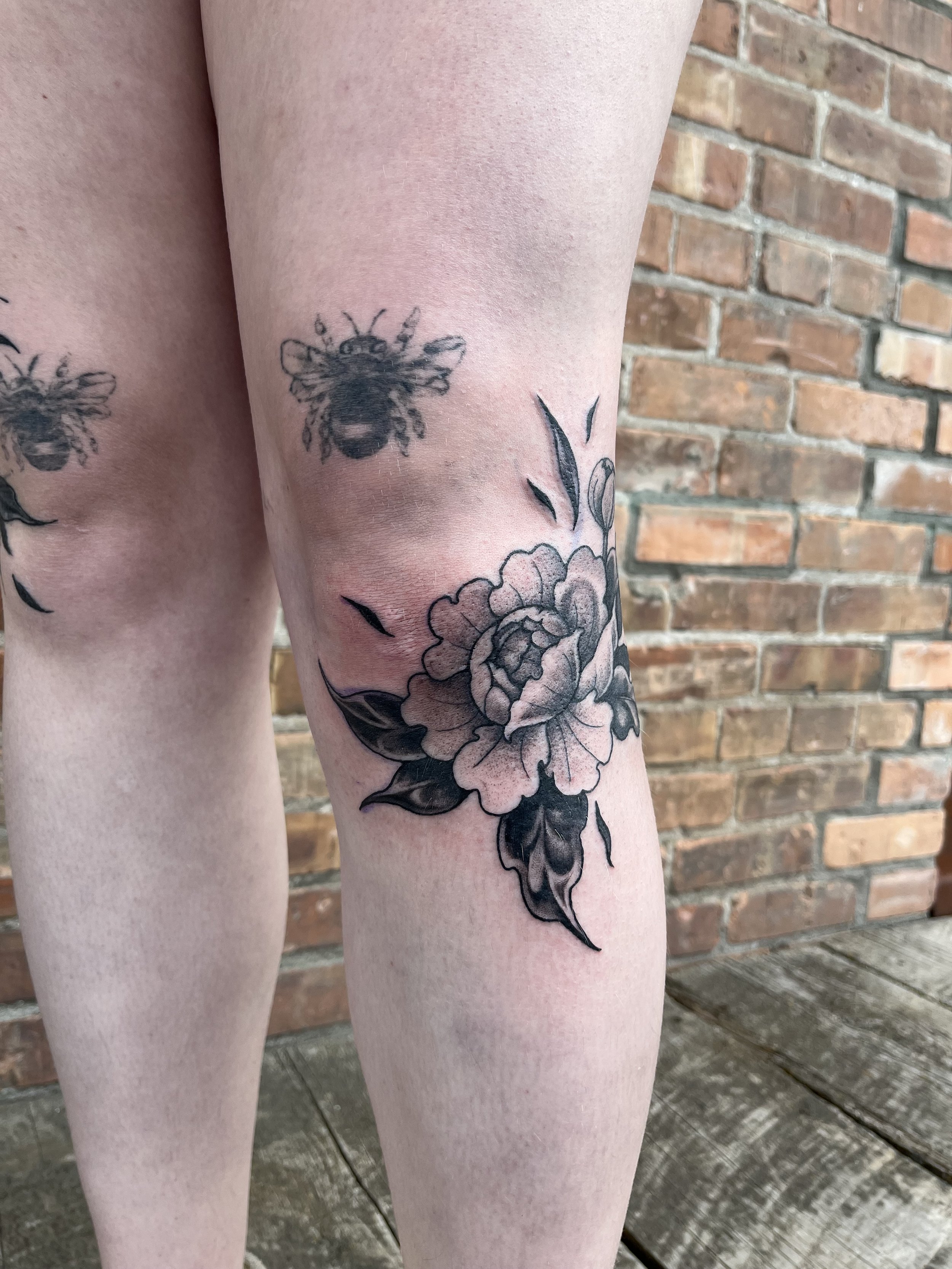 Close-up of a person's legs with floral and bee tattoos on the thigh, in front of a brick wall.