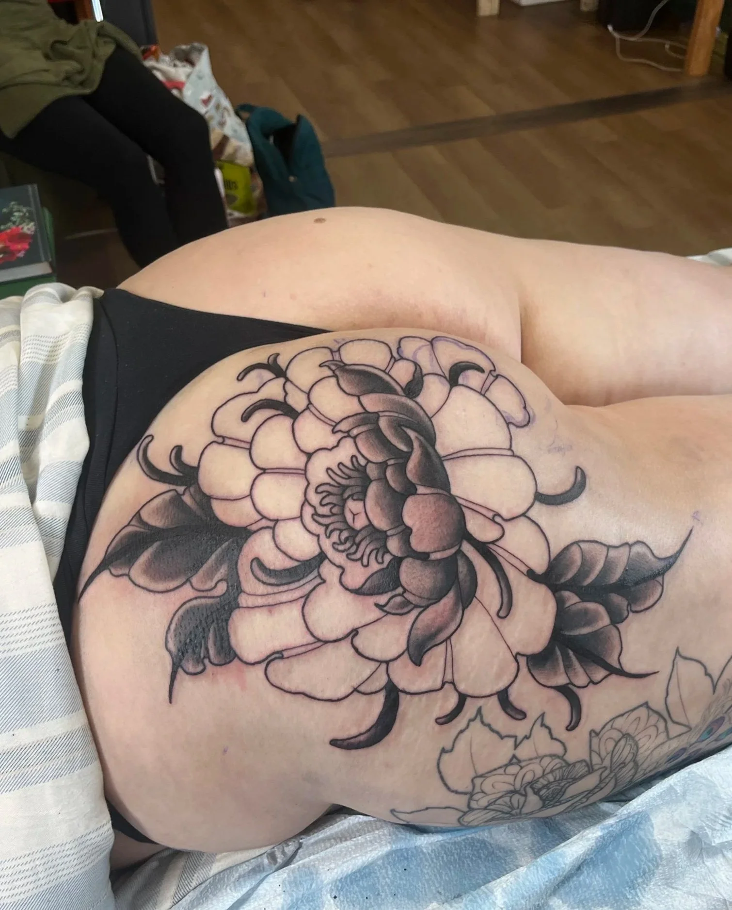Close-up of a person's thigh with a large black and gray floral tattoo, lying on a bed, with a wooden floor in the background and various objects around.