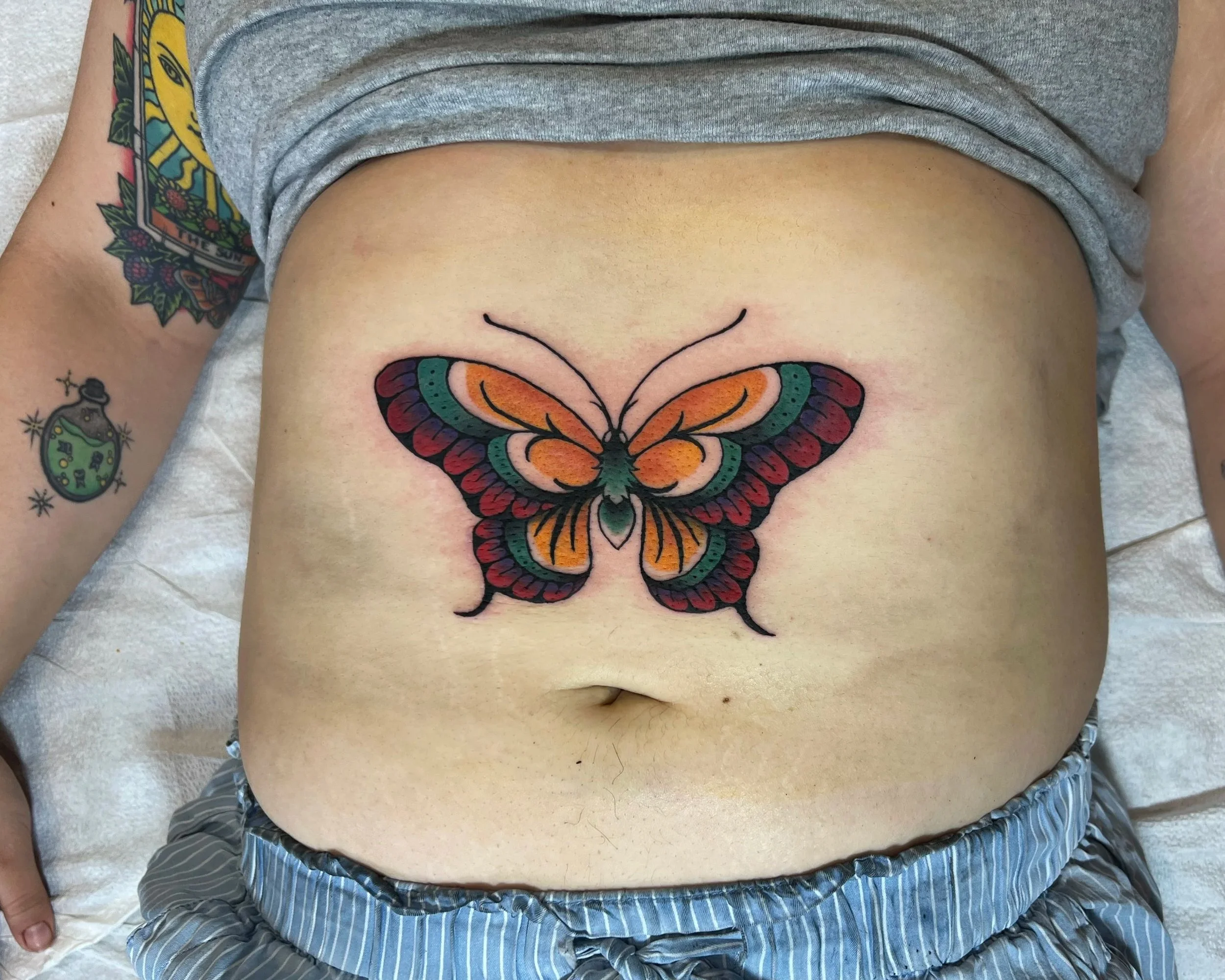A person lying on a bed with a colorful butterfly tattoo on their chest and tattoos on their right arm, wearing a gray T-shirt and striped shorts.