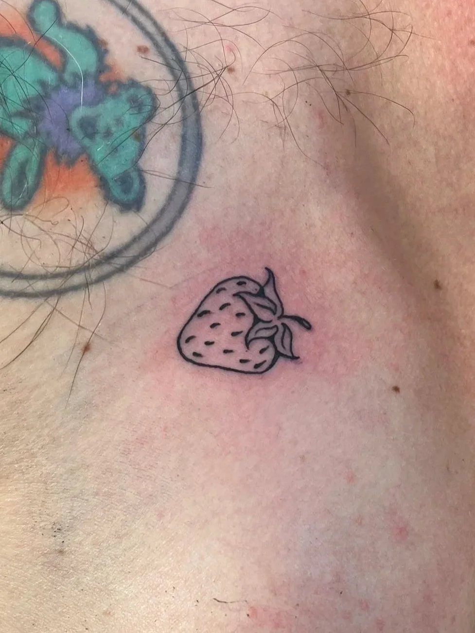 A small strawberry tattoo with simple black outline on skin, near a colorful tattoo and body hair.