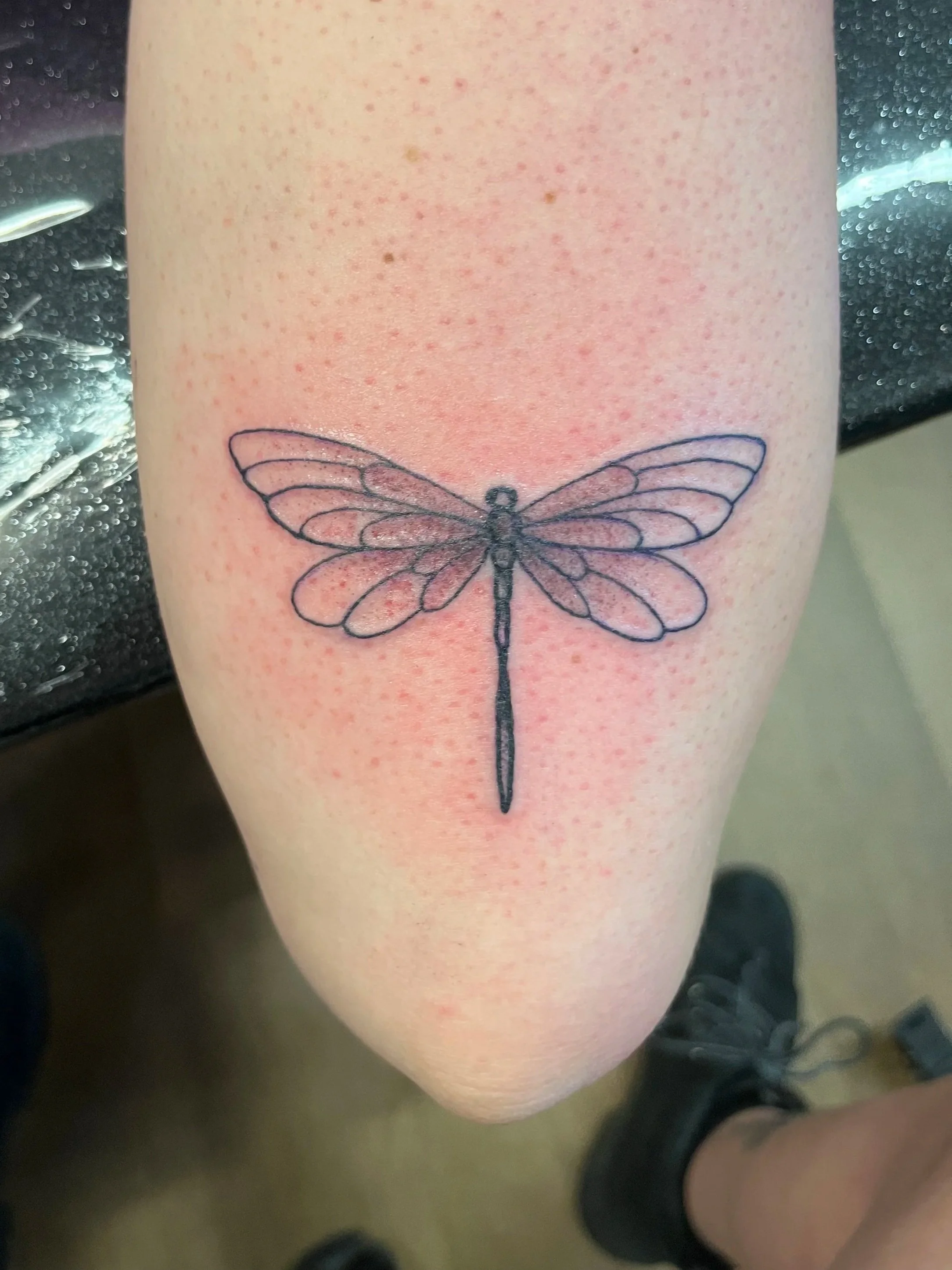 Close-up view of a person's thigh with a black outline tattoo of a dragonfly. The skin around the tattoo appears slightly red and irritated.