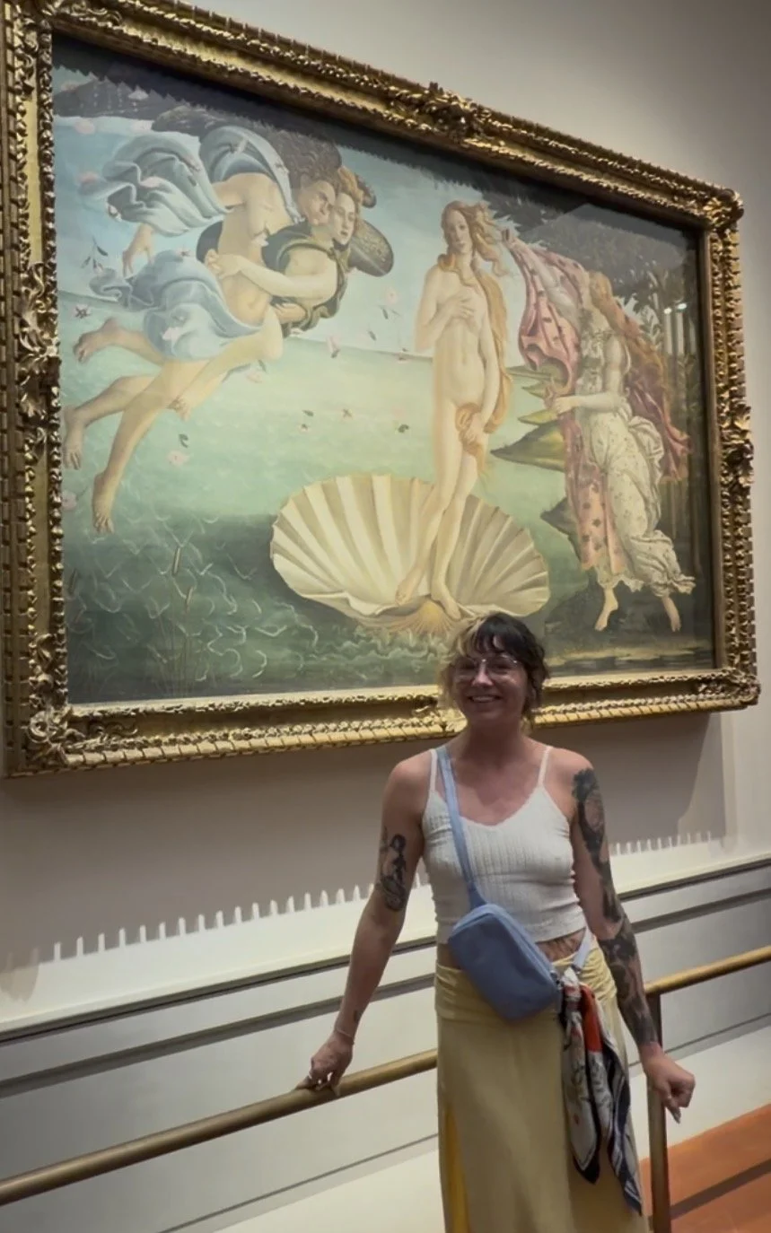 A woman with tattoos on her arms, wearing a white tank top and yellow skirt, standing in front of a large framed painting, smiling in an art gallery.
