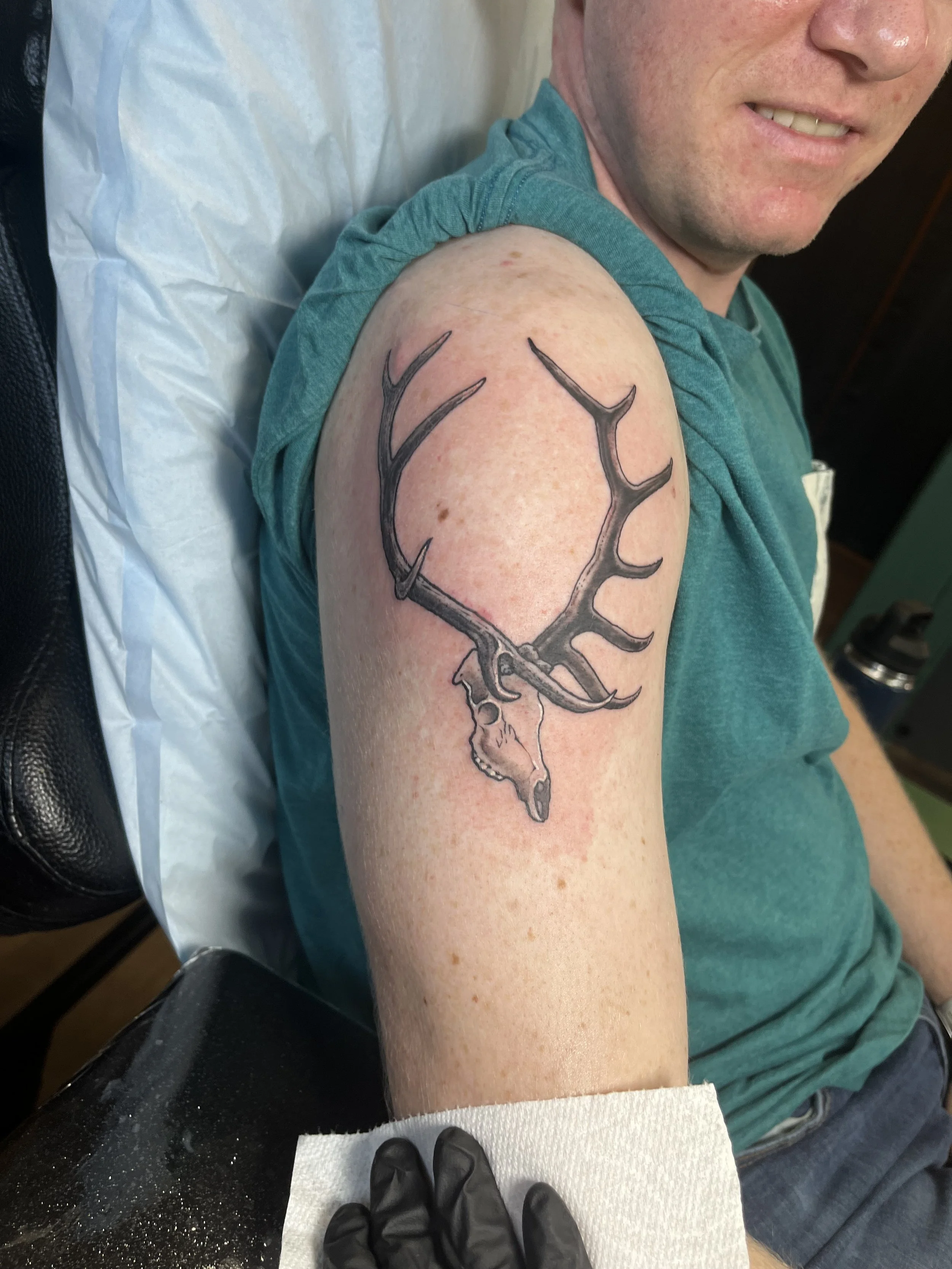 A man sitting in a tattoo chair with a fresh black ink deer antler tattoo on his upper arm, a person wearing black gloves and black pants is preparing to work on the tattoo, a white paper towel is placed on the black surface of the tattoo station, an