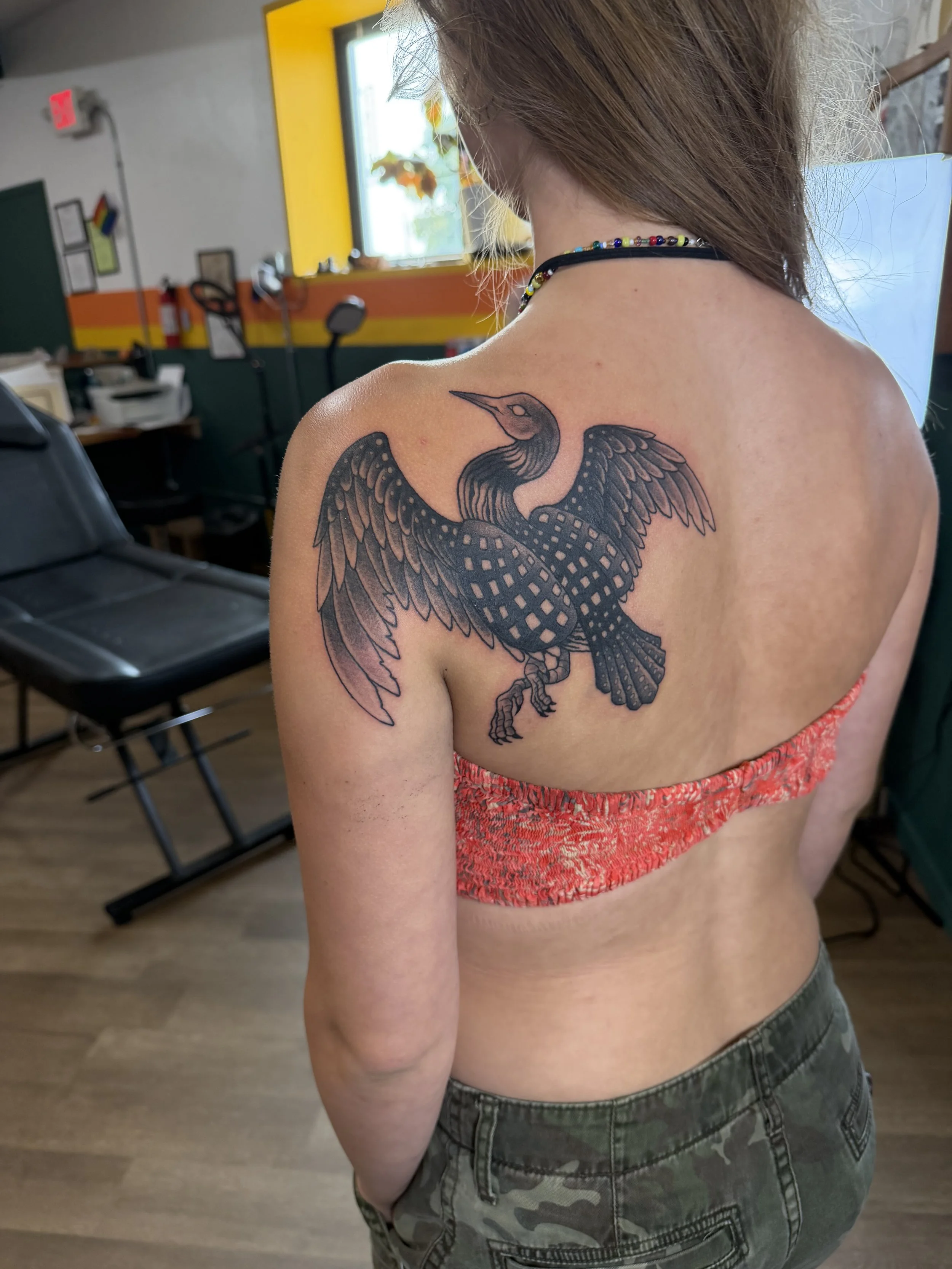 A woman with a colorful beaded necklace displaying a tattoo of a stylized bird on her shoulder, in an indoor setting with furniture and a window in the background.