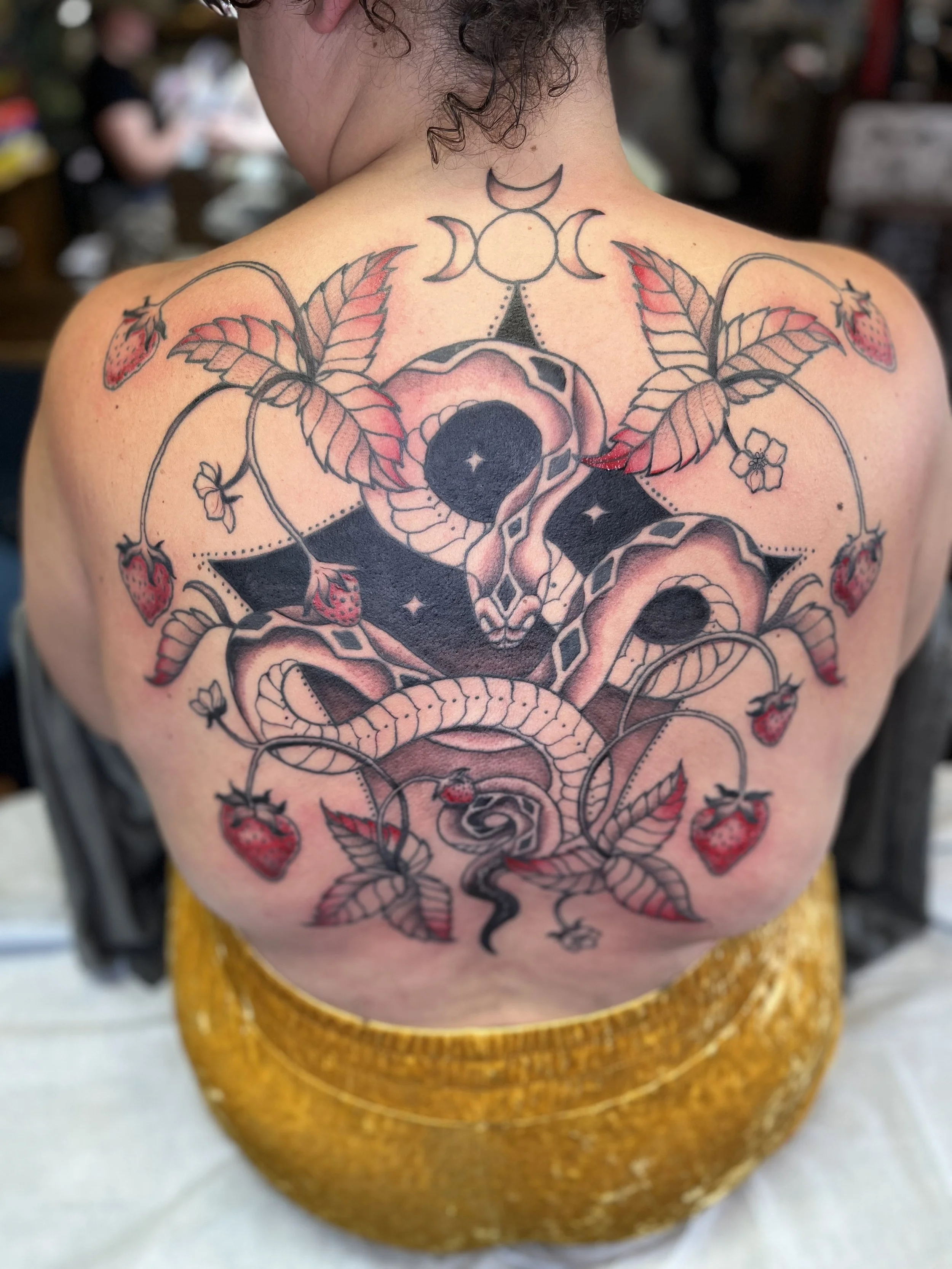 Colorful tattoo on a person's back featuring a snake coiled around a rose with detailed leaves, strawberry elements, and crescent moon symbols above.