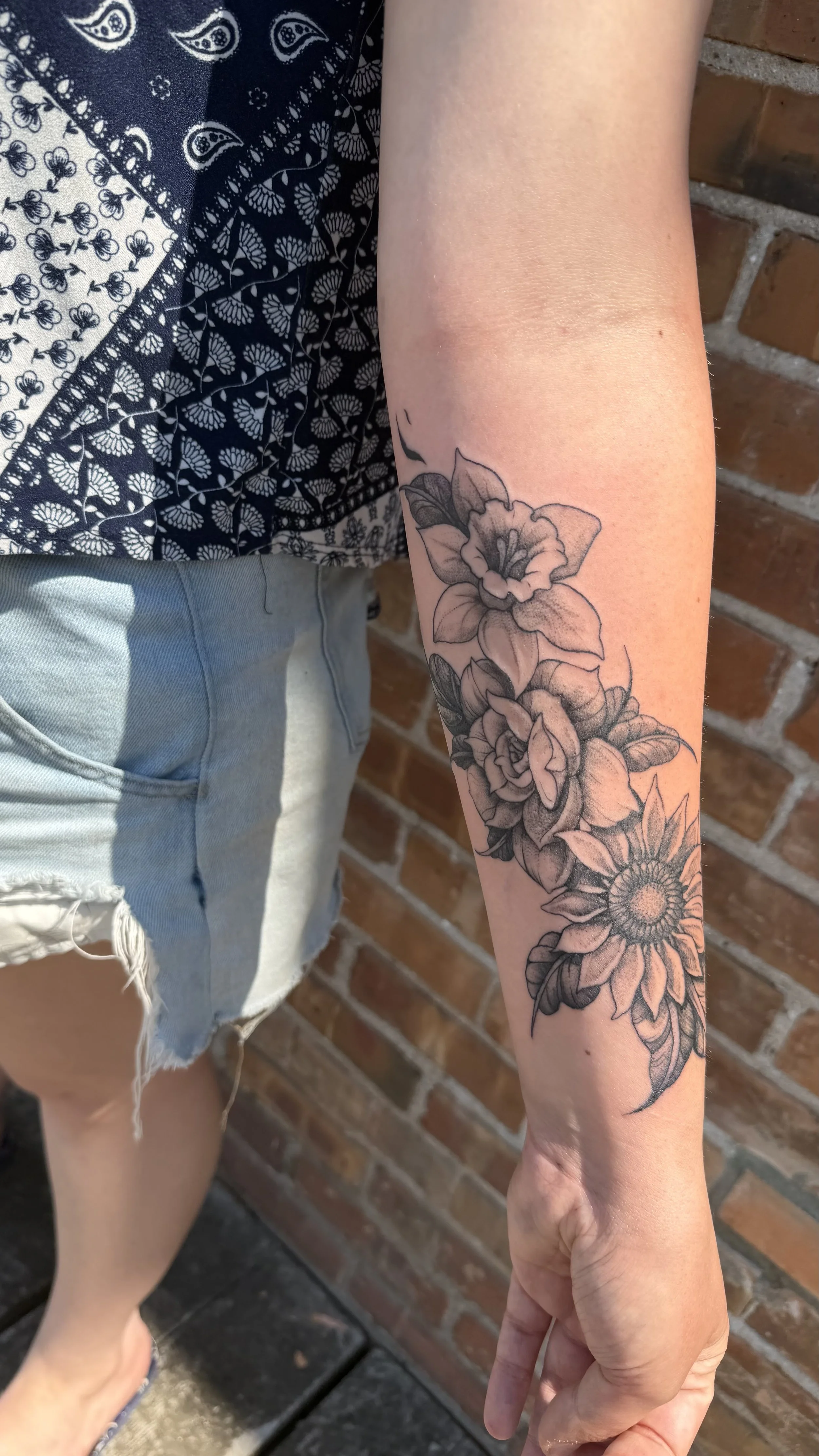 A person's arm with a black and grey floral tattoo featuring flowers and leaves, against a brick wall background.