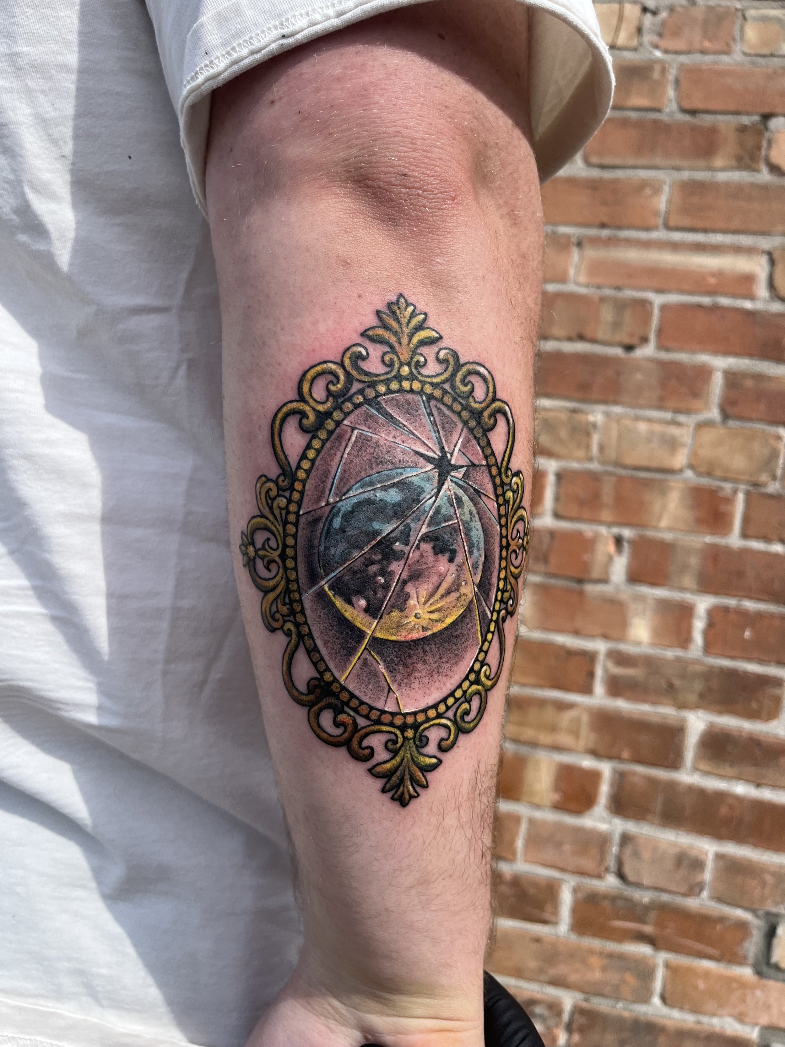 A detailed, colorful tattoo of a broken circular mirror with a gold ornate frame is on a person's inner forearm. The background features a brick wall.