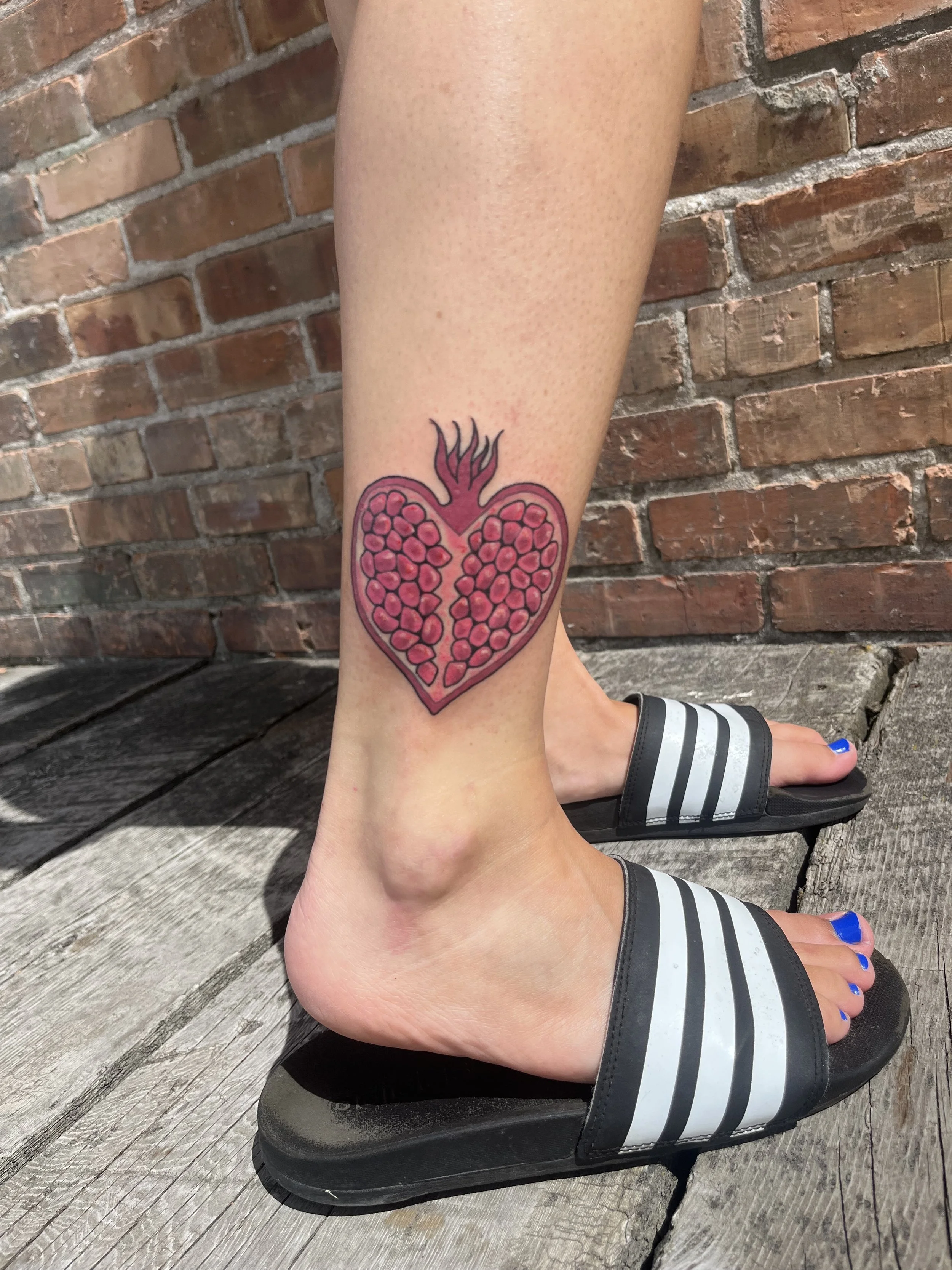 A person's lower leg with a pomegranate tattoo above the ankle, wearing black and white slide sandals, standing on a wooden surface in front of a brick wall.