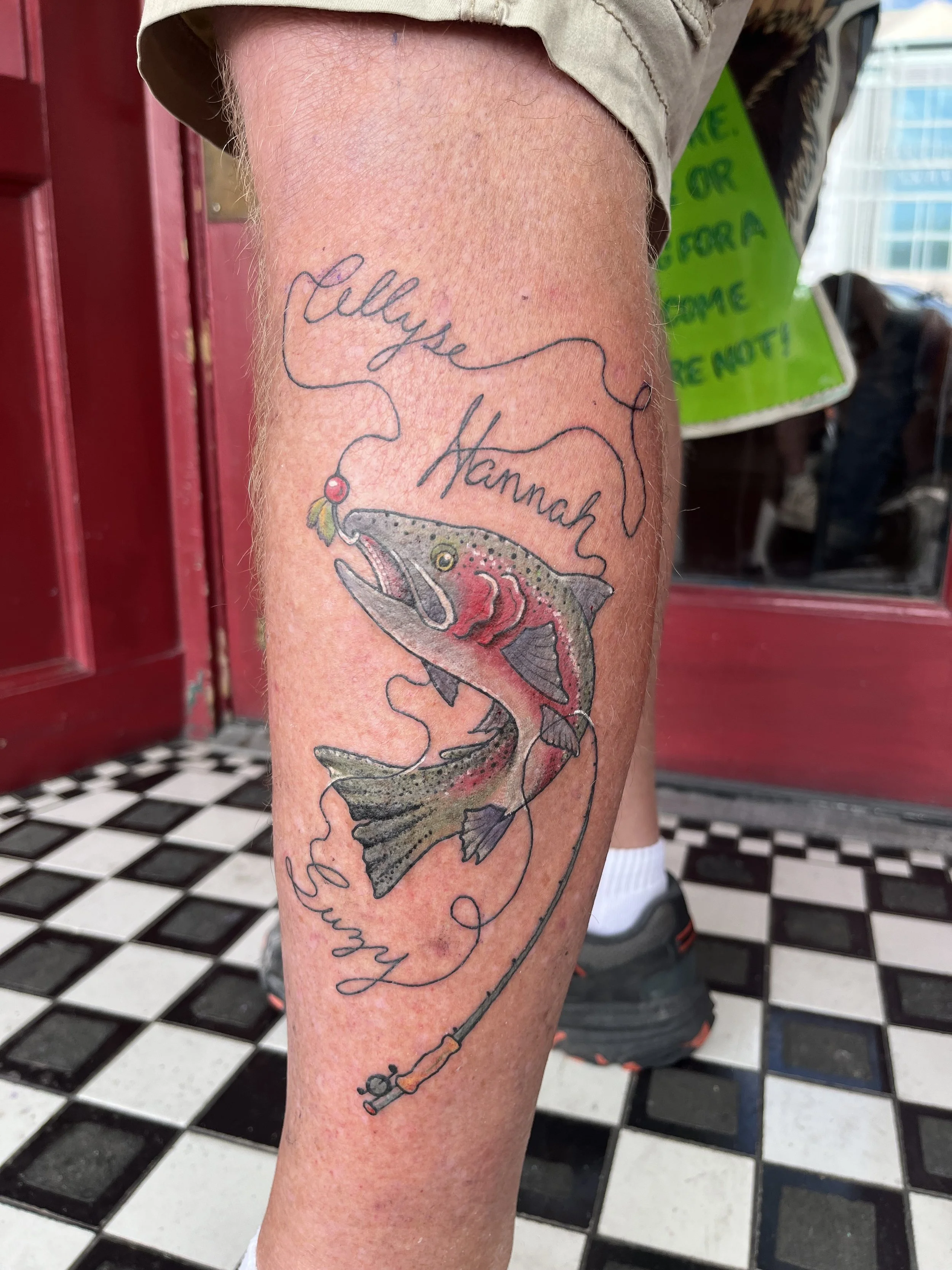 A tattoo on a person's calf of a detailed fish leaping out of water, with the words "Lillys Hannah" written above and below in cursive.