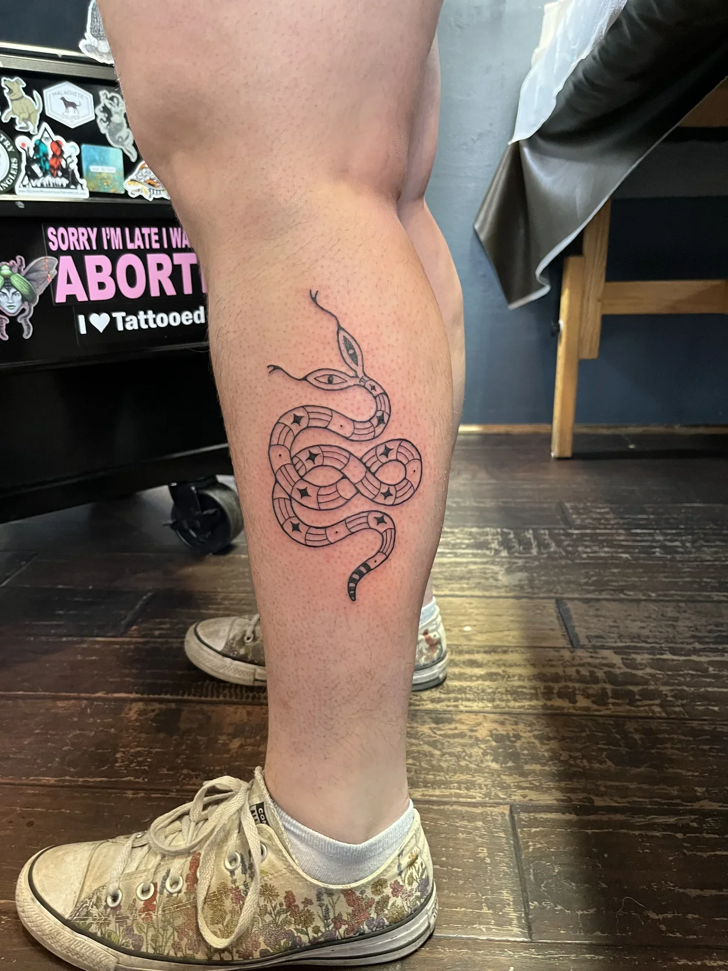 A person's leg with a tattoo of a snake with multiple eyes, standing on a wooden floor. The person is wearing a beige sneaker with a floral pattern, white socks, and shorts. A sticker display with the text 'Sorry I'm late I was ABORTED' and 'I ❤️ Tat