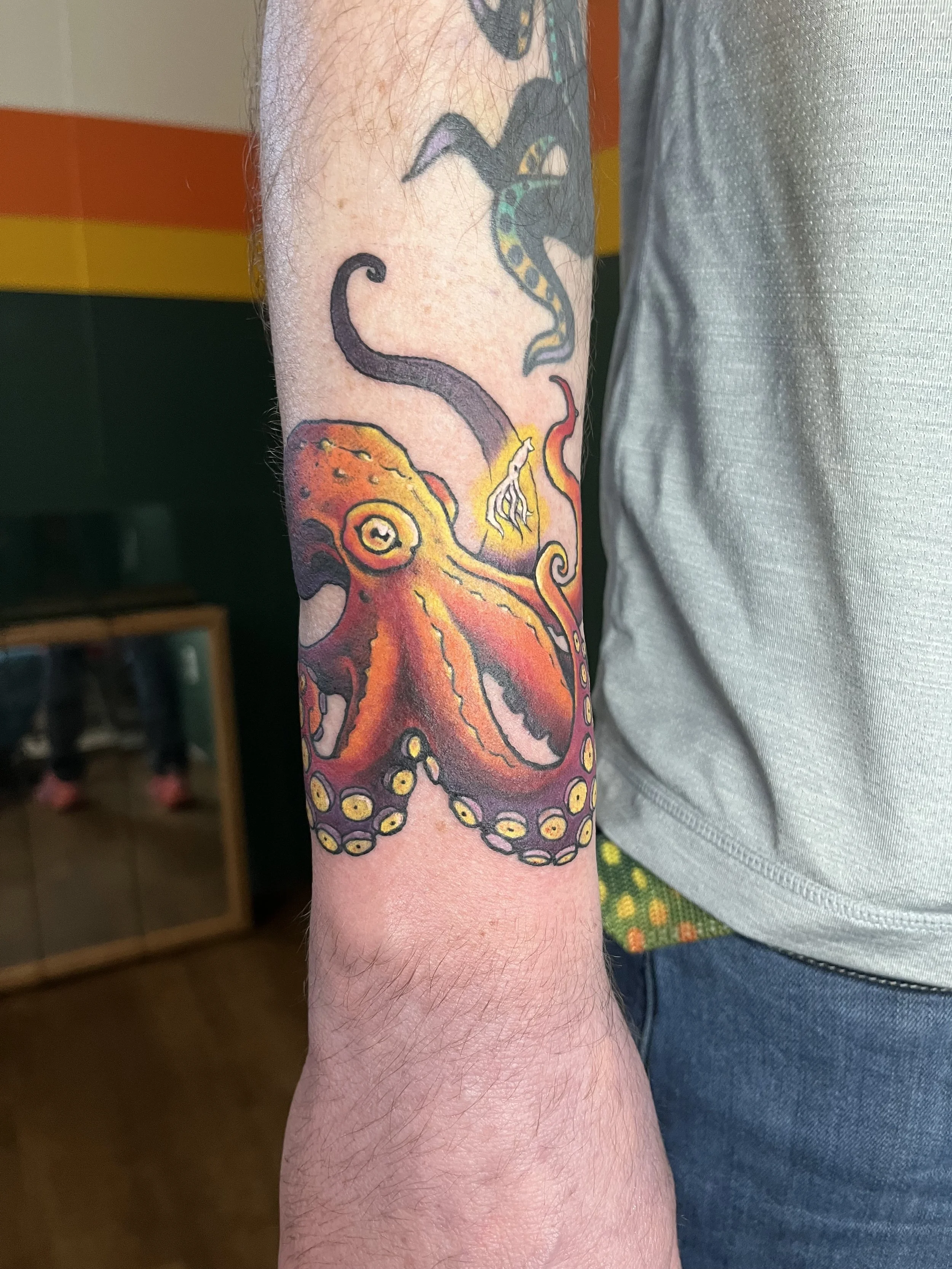 Colorful octopus tattoo on person's forearm, with a background of orange and yellow stripes.