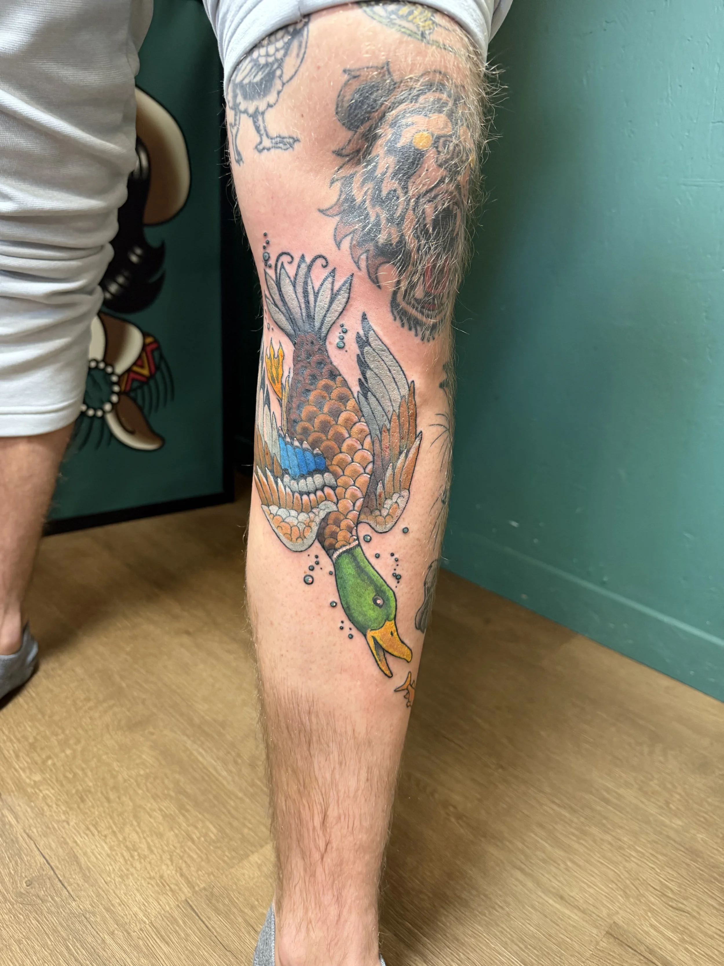 Tattoo of a lion's head, a duck, and a floral design on a person's leg.