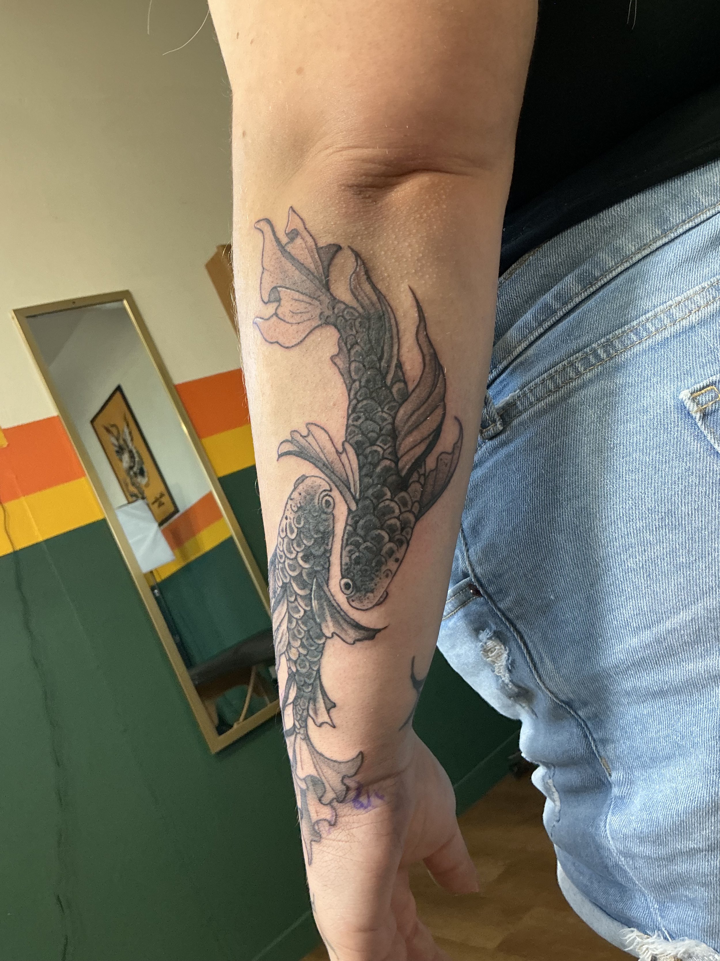 A tattoo of a koi fish on a person's forearm, with the fish swimming upward and detailed scales and fins, in black and gray ink.