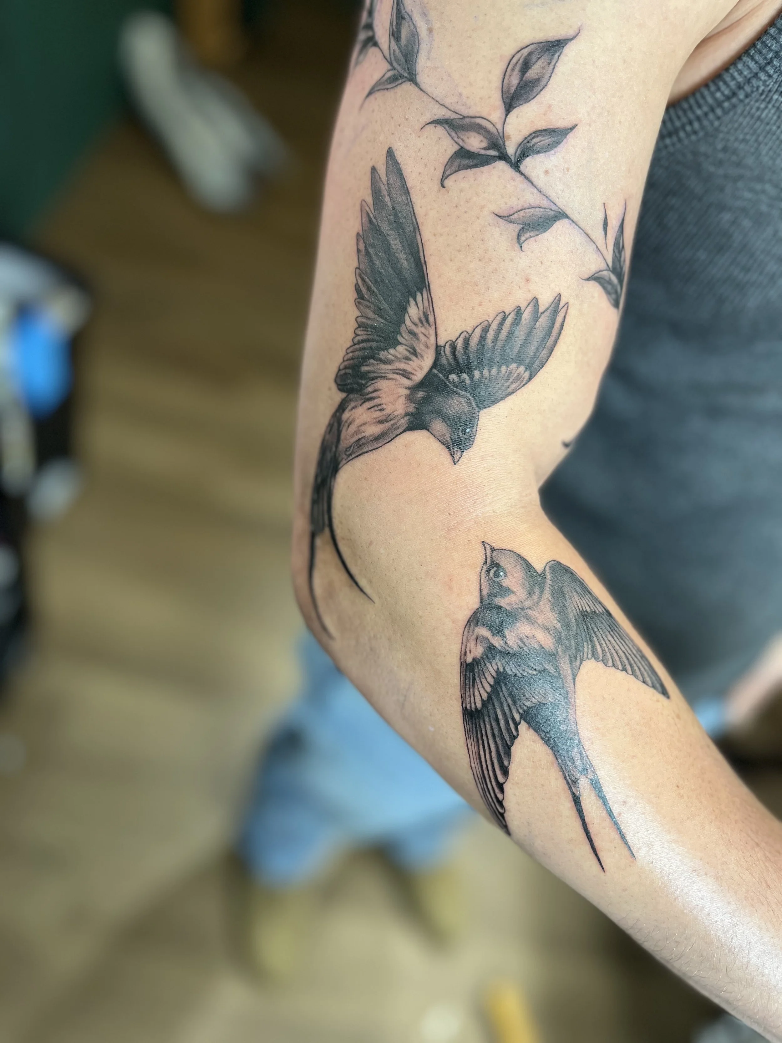A black and gray tattoo of two birds flying, one larger and one smaller, on a person's arm.
