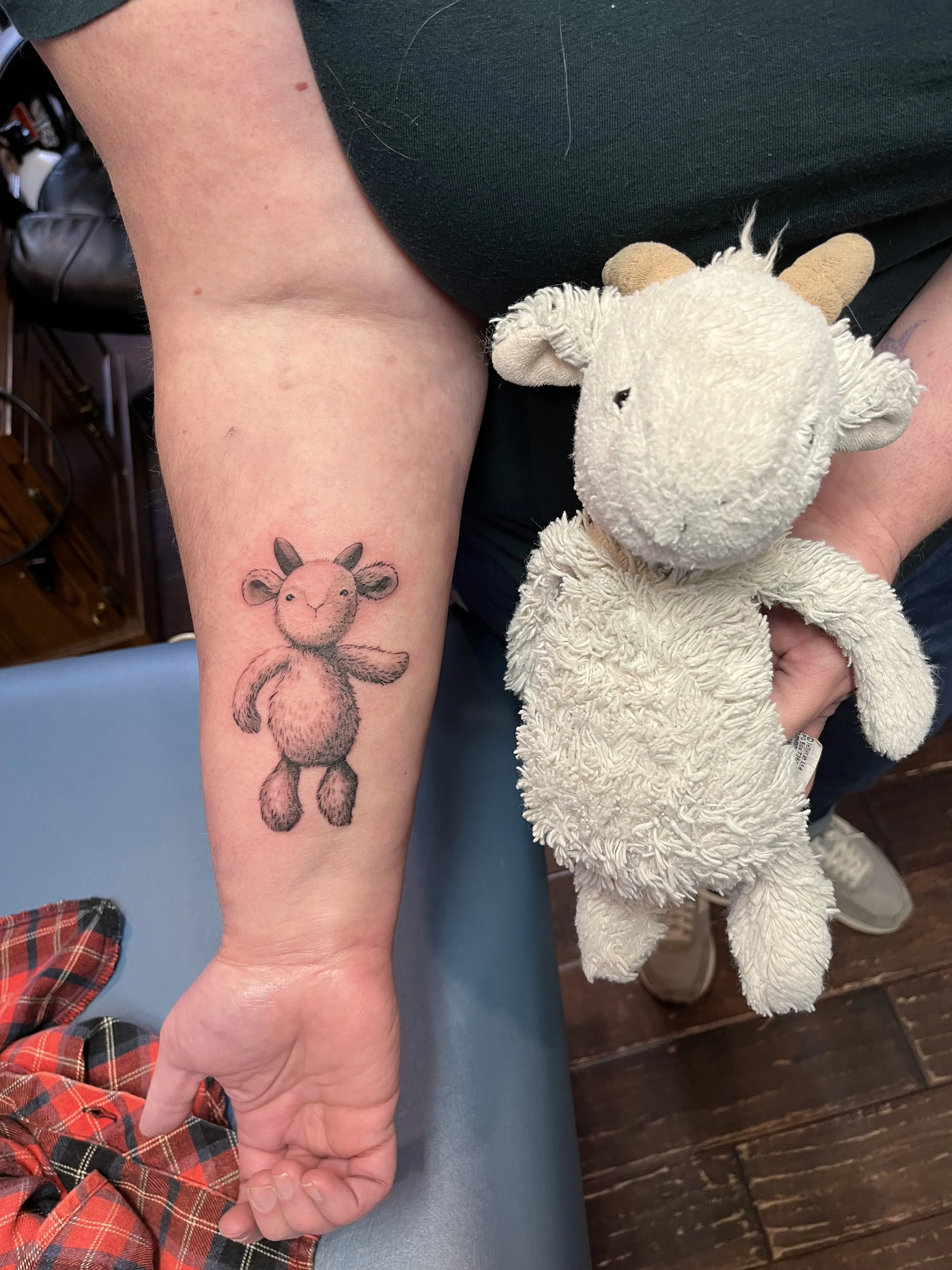 Old worn plush teddy bear being held by a person with a small cartoon style fox tattoo on their forearm, sitting in a room with wooden floors.