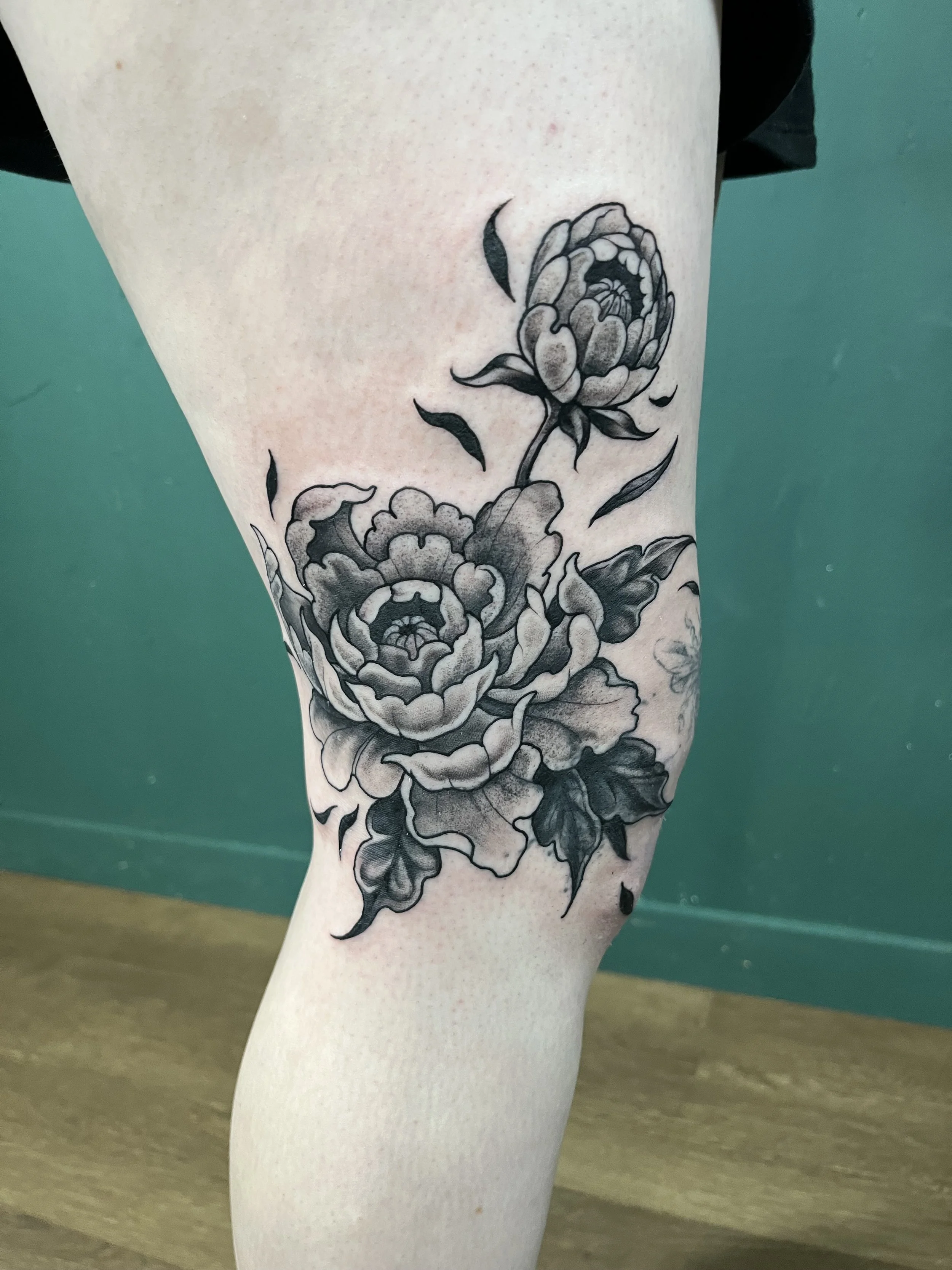 A detailed black and gray floral tattoo featuring large peony flowers on a person's thigh.