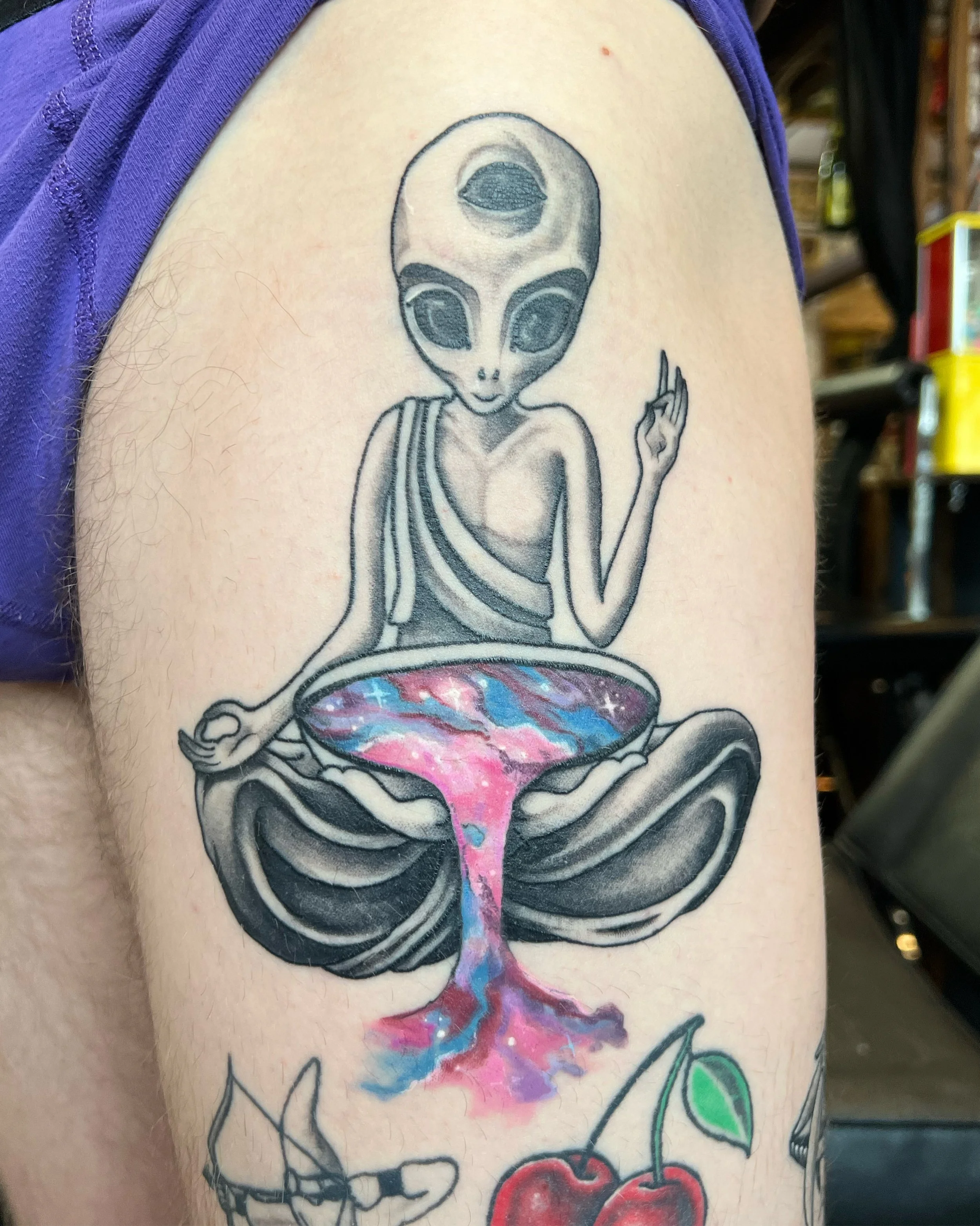 Tattoo of an alien sitting cross-legged with a large head and big black eyes, holding a bowl of colorful galaxy-like liquid, on a person's upper arm.