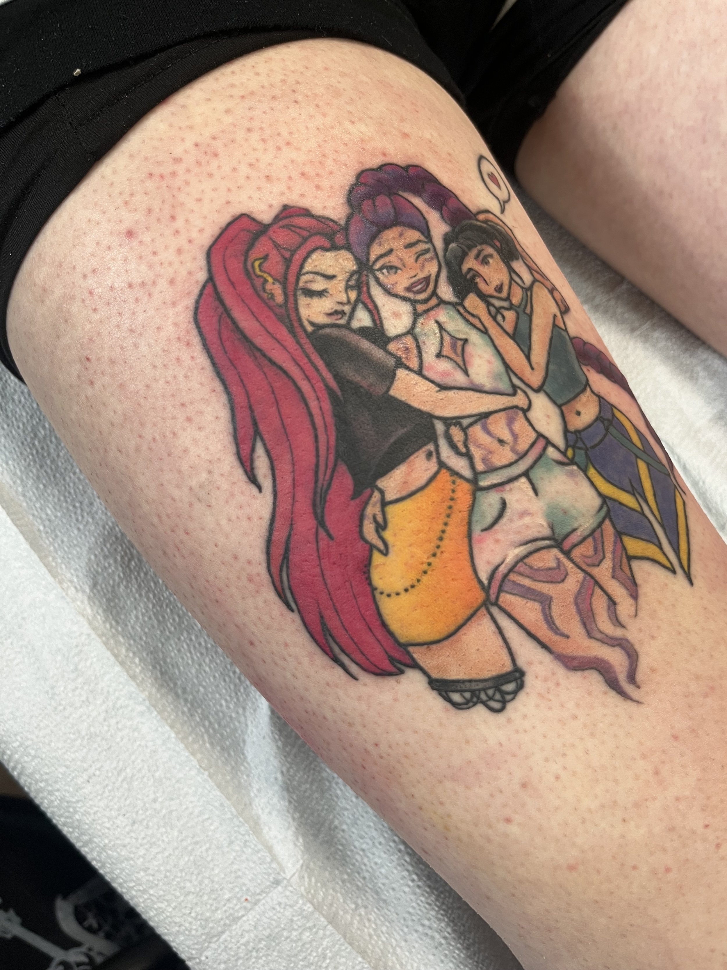 A colorful tattoo of three stylized girls with different hair colors and styles, posing together with a cheerful expression. One has long red hair, another has purple braids, and the third has short black hair. The tattoo features bold outlines and b