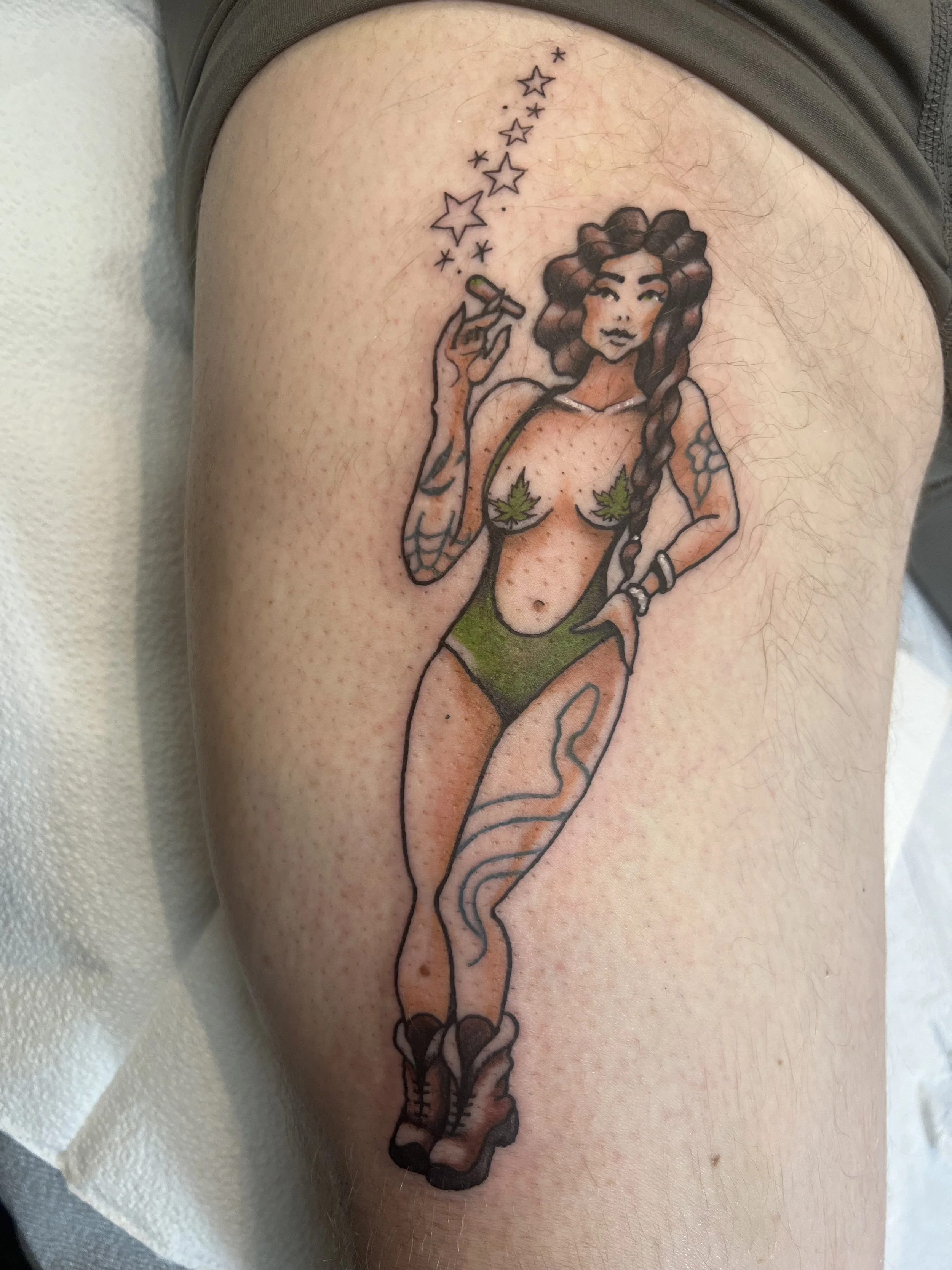 A tattoo of a woman with curly hair, holding a bong with stars emanating from it, wearing a green outfit with leaf pasties, and standing on a pair of sneakers.