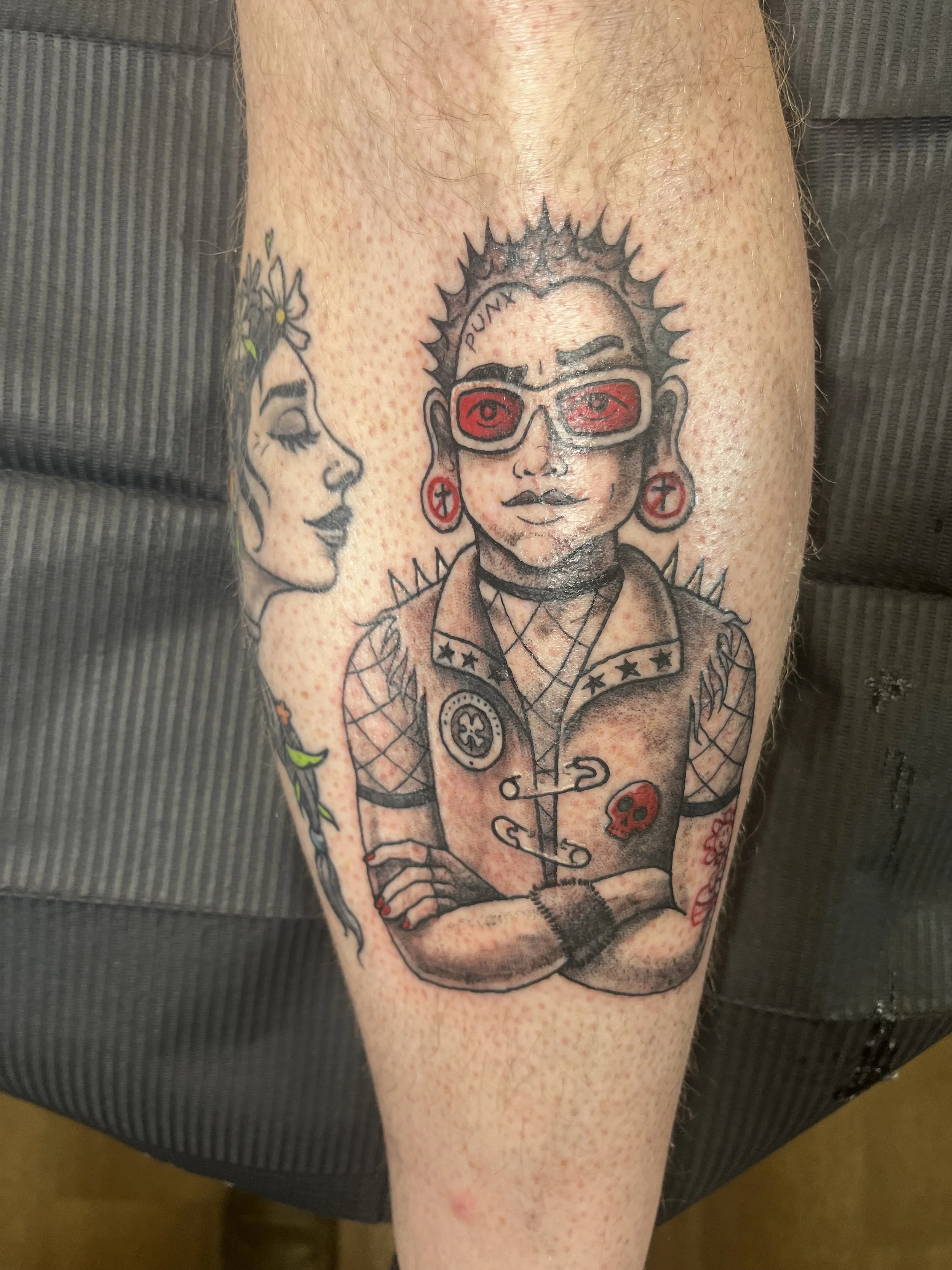 Tattoo of a woman wearing glasses with the word 'PUNK' on her forehead, earrings with crosses, and spiked hair. Another face of a woman with flowers in her hair is partially visible.