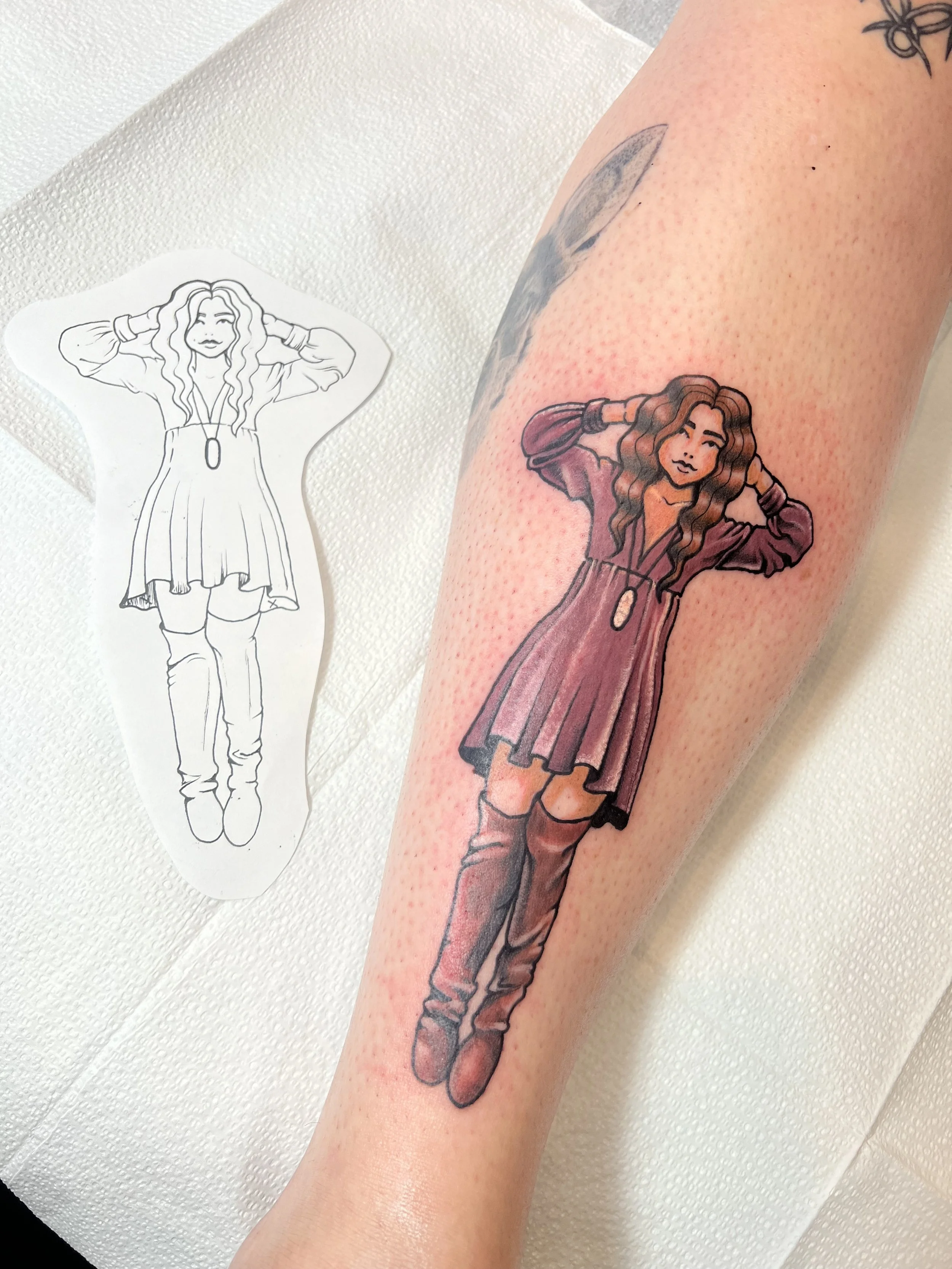 Colorful tattoo of a woman with long hair, wearing a jacket and boots, on a person's arm, with a tattoo outline sketch of the same design beside it.