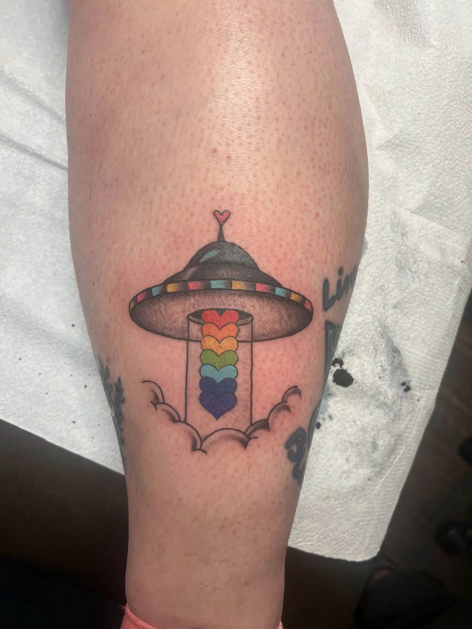 A tattoo of a lamp with a rainbow-colored rainbow pouring from it, with a small pink heart at the top of the rainbow. The tattoo is on a person's arm.
