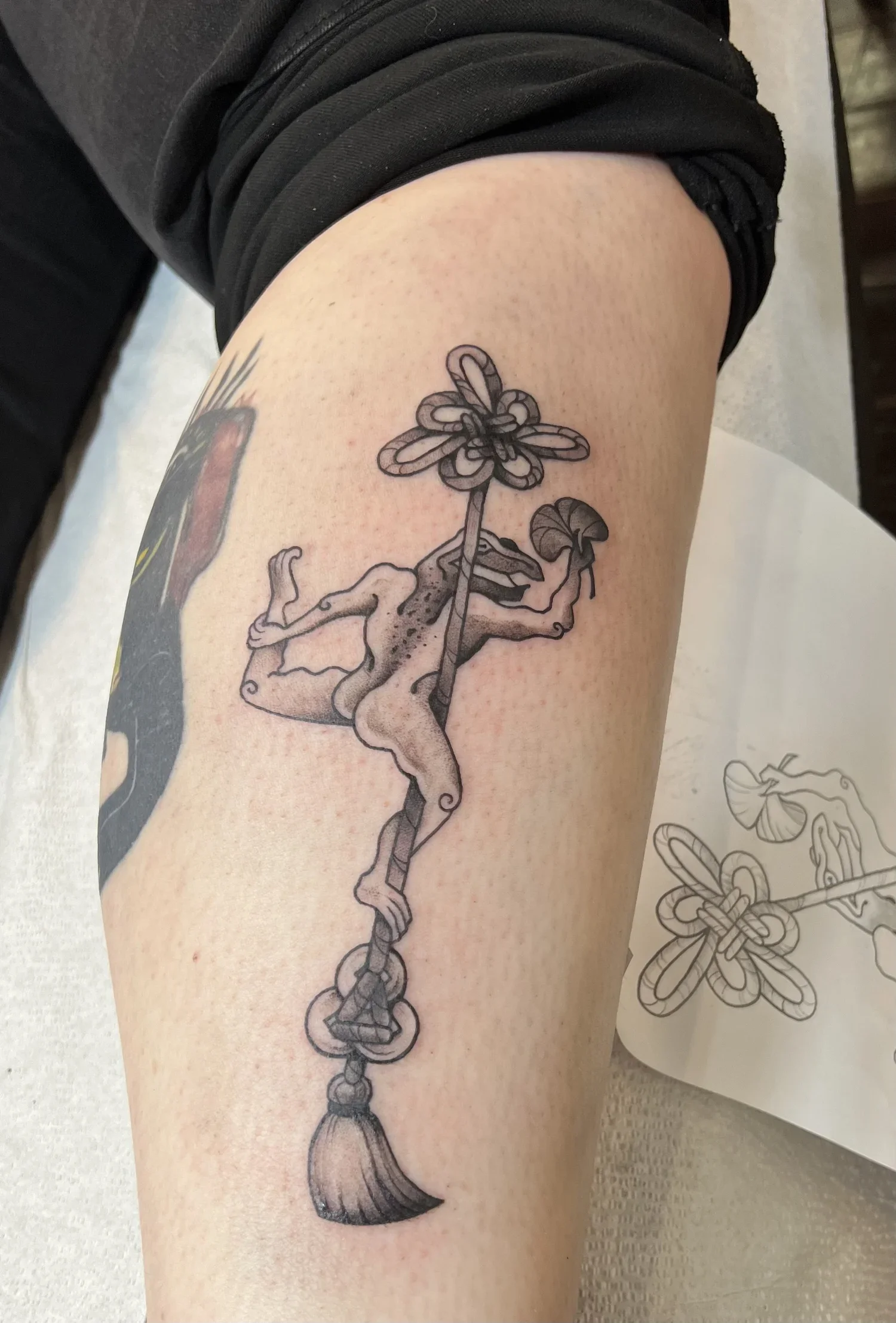 Tattoo of a unicorn with a flower-shaped horn, standing on its hind legs, connected to a chain with a loop at the end, on a person's upper arm.