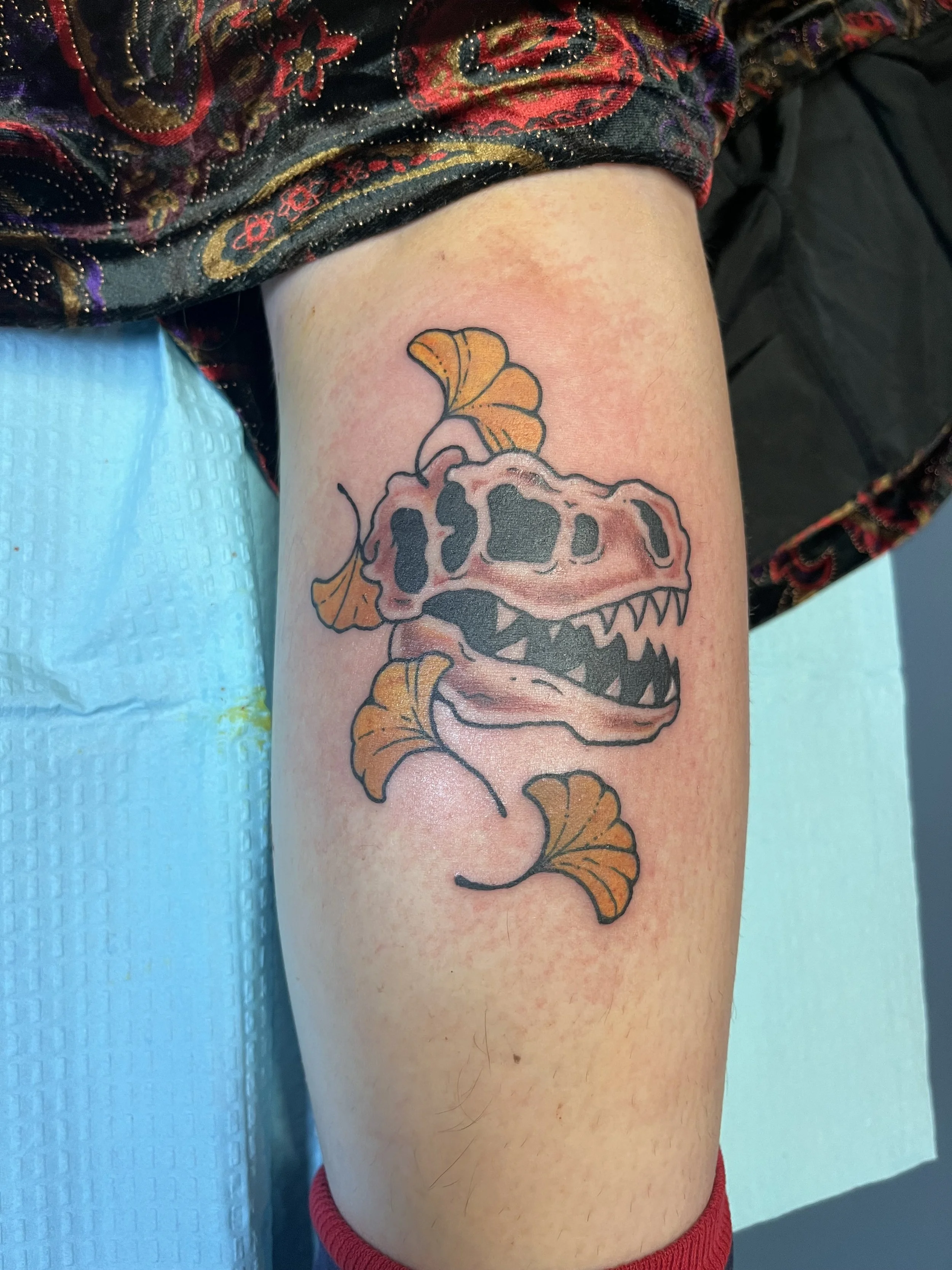 Tattoo of a dinosaur skull with orange ginkgo leaves on a person's upper arm.