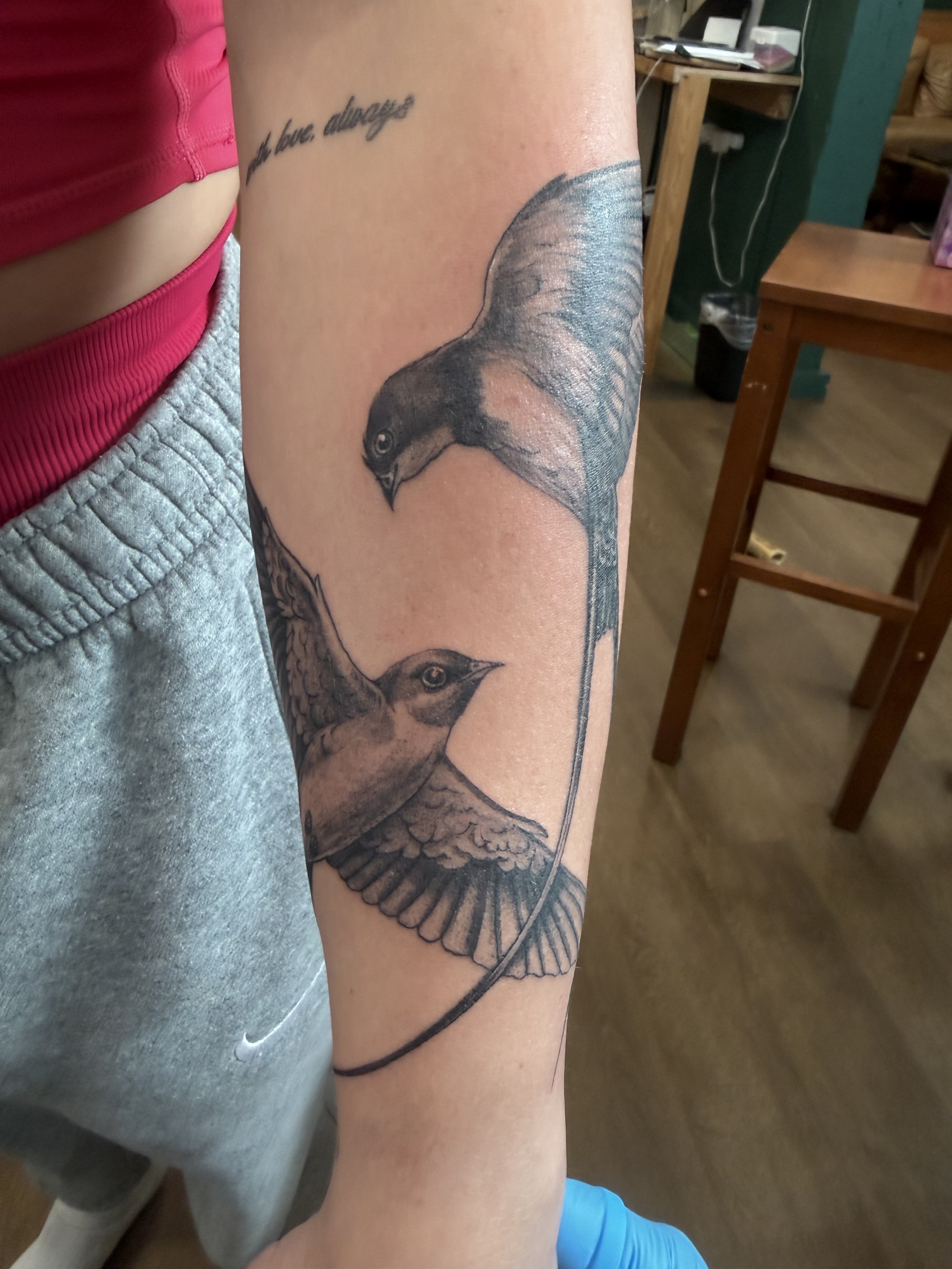 Close-up of a person's upper arm with a detailed black and gray tattoo of a bird, possibly a hawk or falcon, with text above it that says 'and love always'; the person is wearing a pink top and gray shorts.