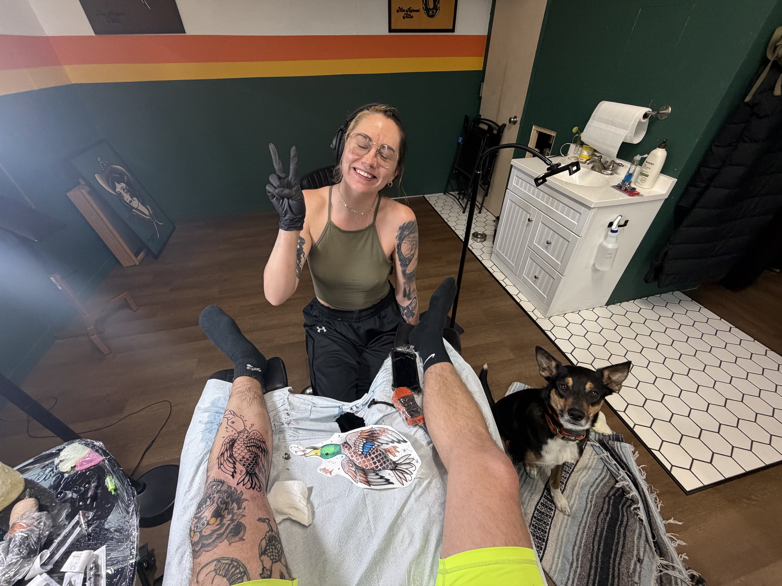 Tattooed person sitting on a tattoo chair at a tattoo studio, with a woman smiling and making a peace sign behind them, and the studio decorated with colorful stripes and artwork on the walls.