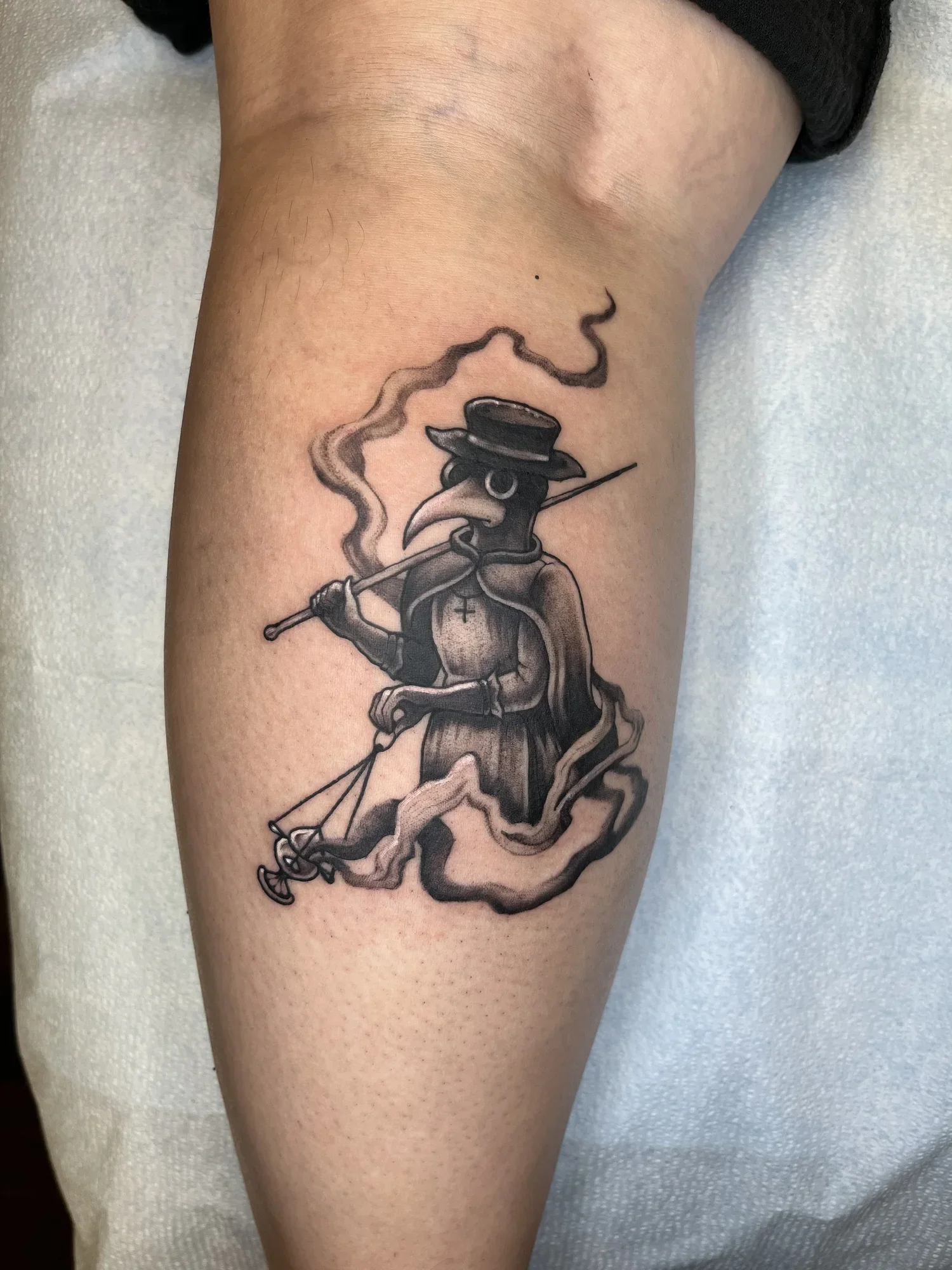 Tattoo of a cartoon bird with a long beak, wearing a wide-brimmed hat, a hoop earring, and an outfit with a cross pendant, holding a thin stick, with a trail of smoke or lines behind it.