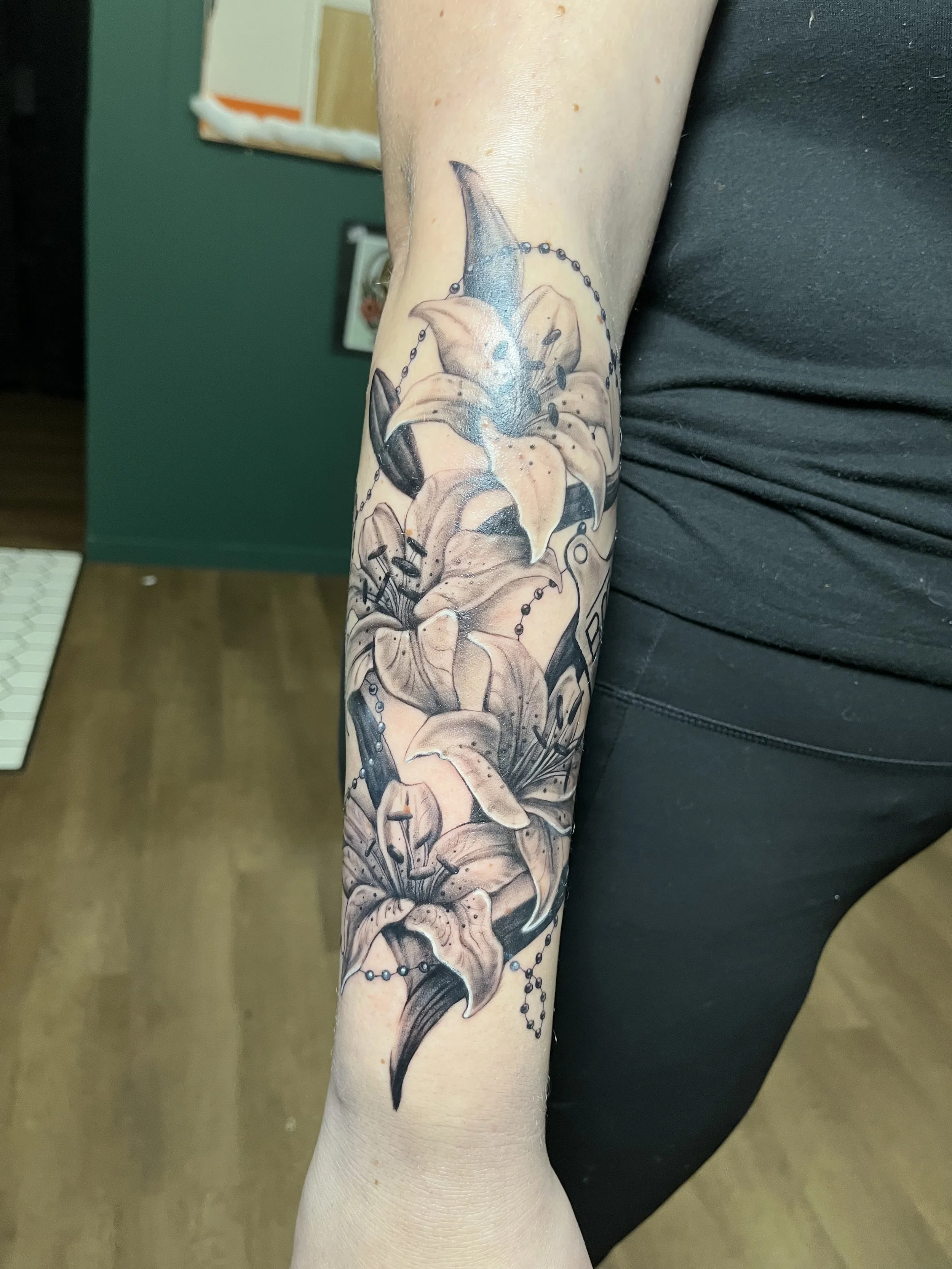 A tattoo of a fully bloomed lily flower on a person's forearm, with detailed black and gray shading and some hint of color on the petals.
