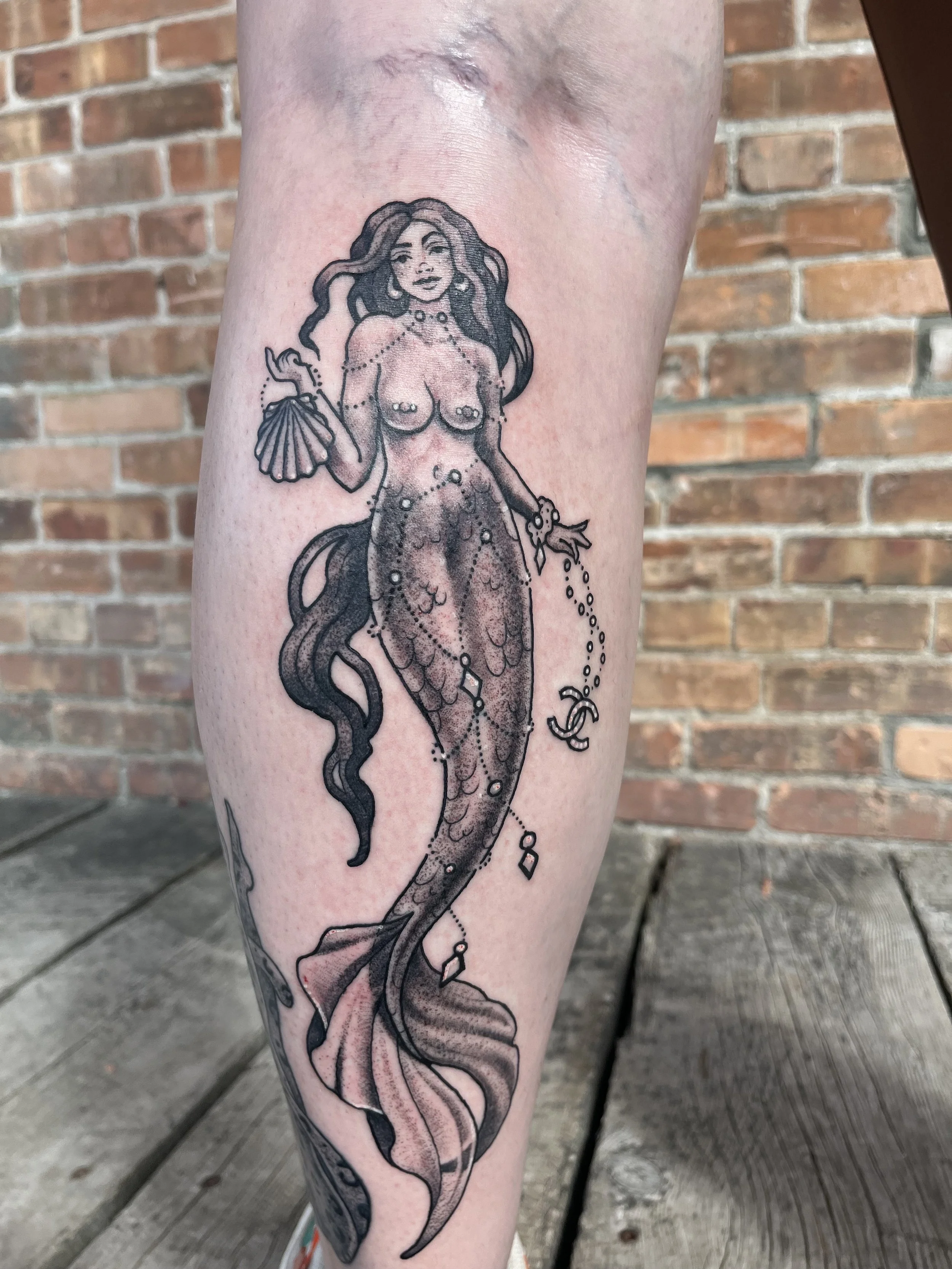Tattoo of a mermaid with long hair, holding a fan and jewelry, with decorative elements around her and a brick wall background.