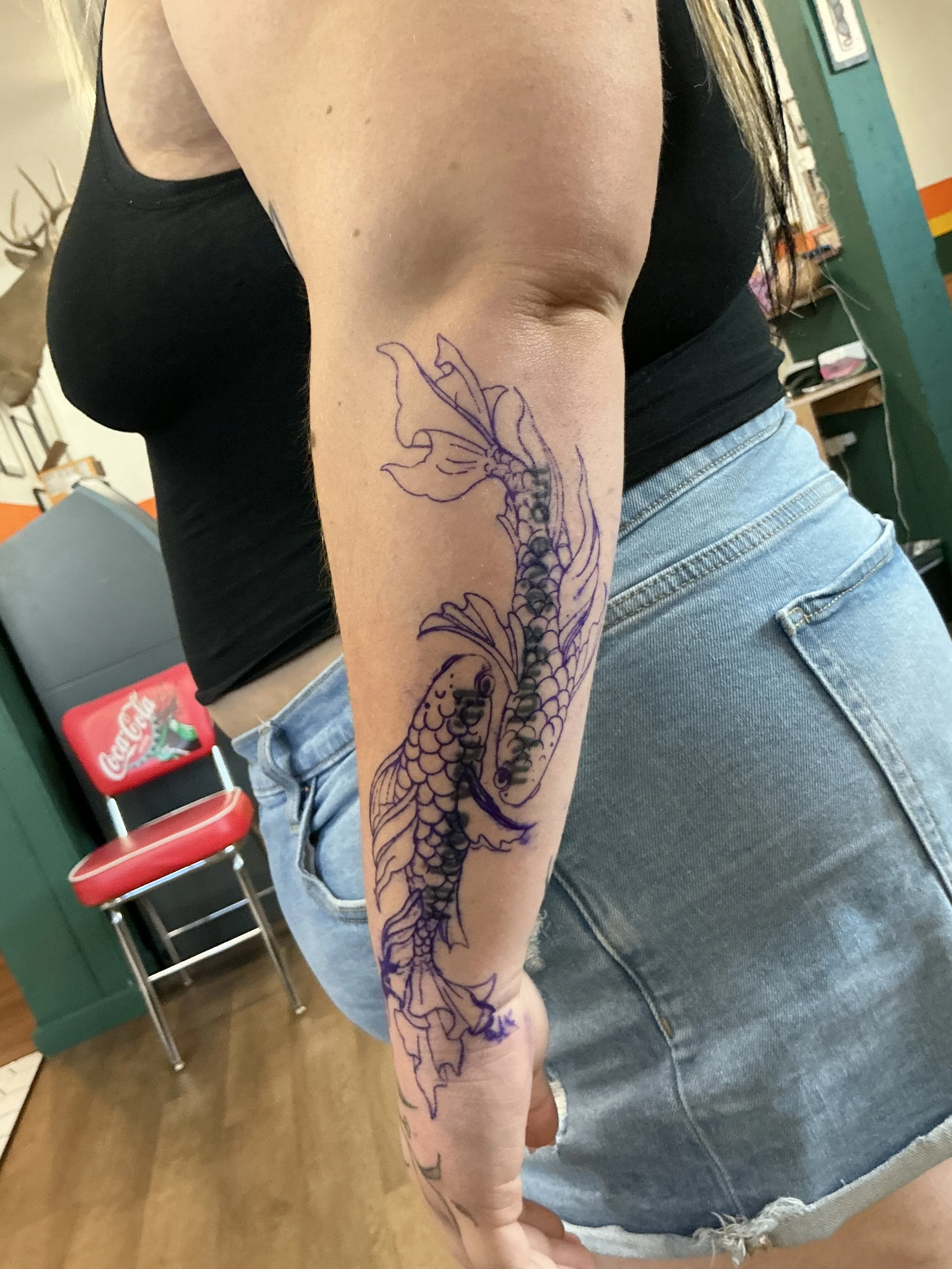 A person's arm with a dragon tattoo drawn in purple ink along the forearm, positioned with a black shirt and blue denim shorts in the background.