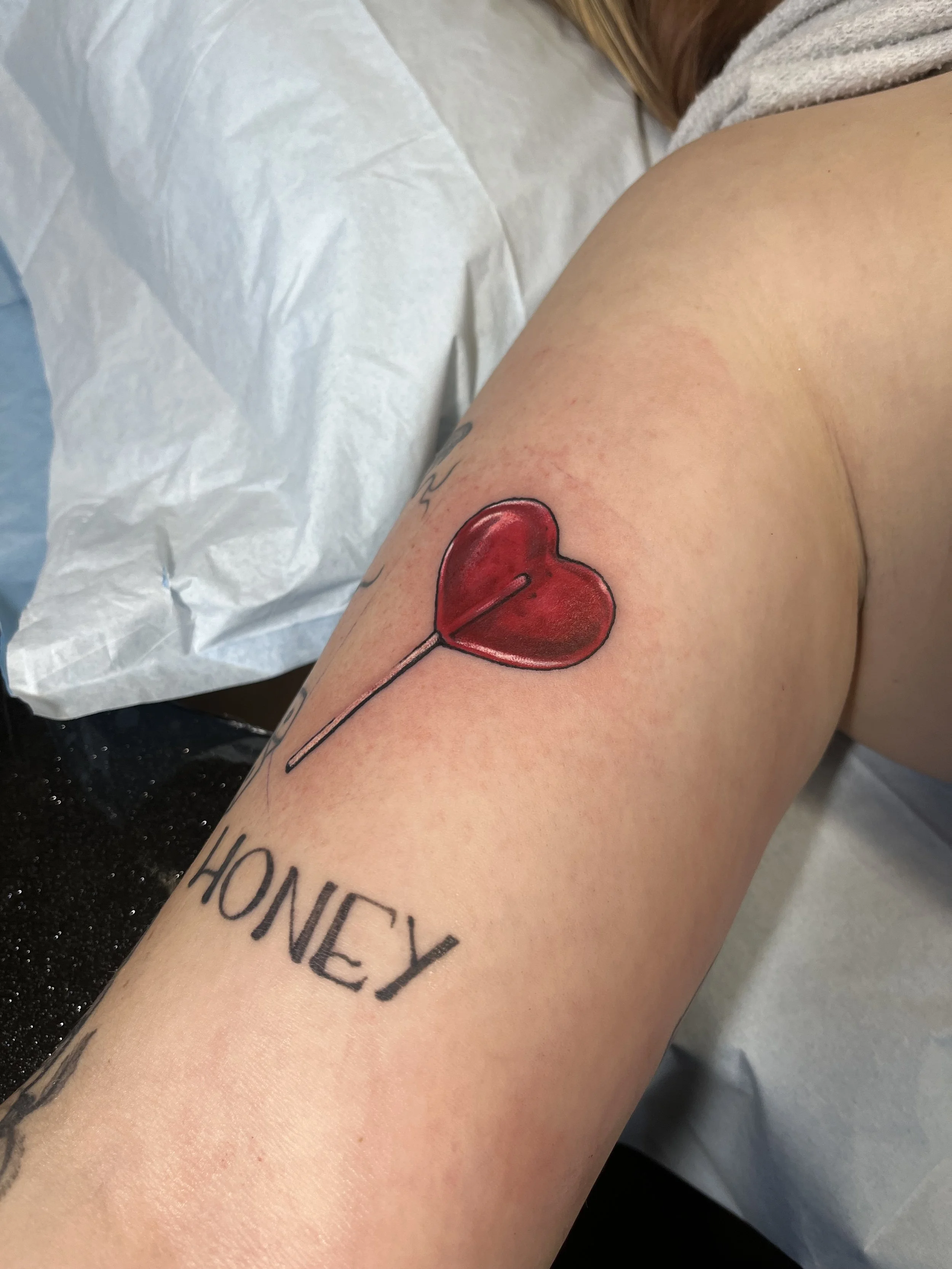 Close-up of a person's arm with a tattoo of a red heart on the upper arm and the word 'HONEY' in black letters on the forearm, lying on a white hospital bed.