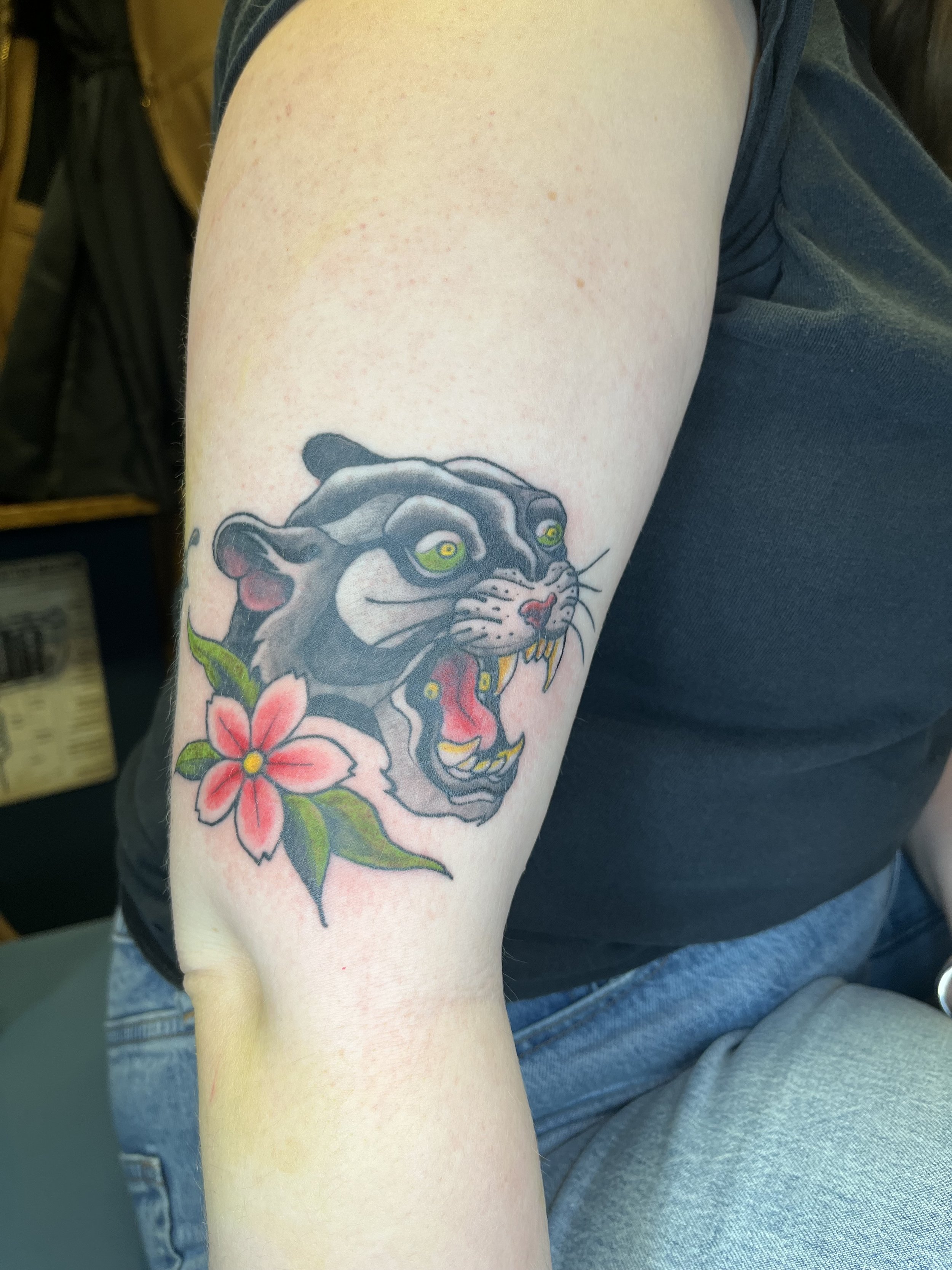 Tattoo of a fierce panther with an open mouth revealing sharp teeth, and a pink flower with green leaves on a person's upper arm.