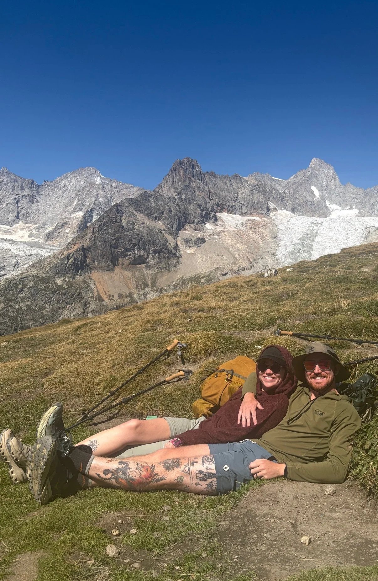 Two smiling hikers lying on the grass in front of a mountain range with rocky peaks and ice patches, surrounded by green grass and a clear blue sky.
