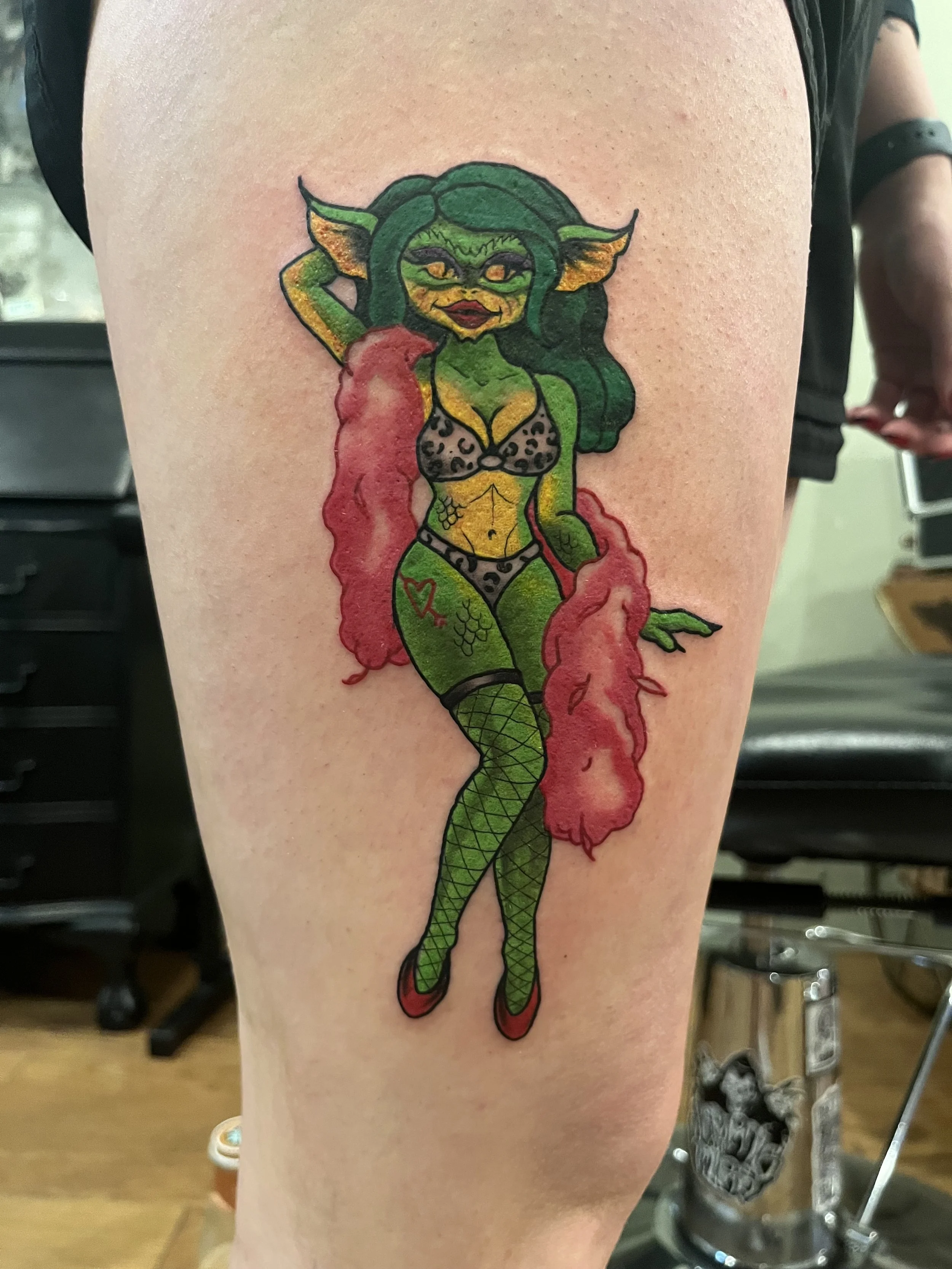 Colorful tattoo of a cartoonish green demon woman with long green hair, leopard print clothing, and red smoky effects around her legs.
