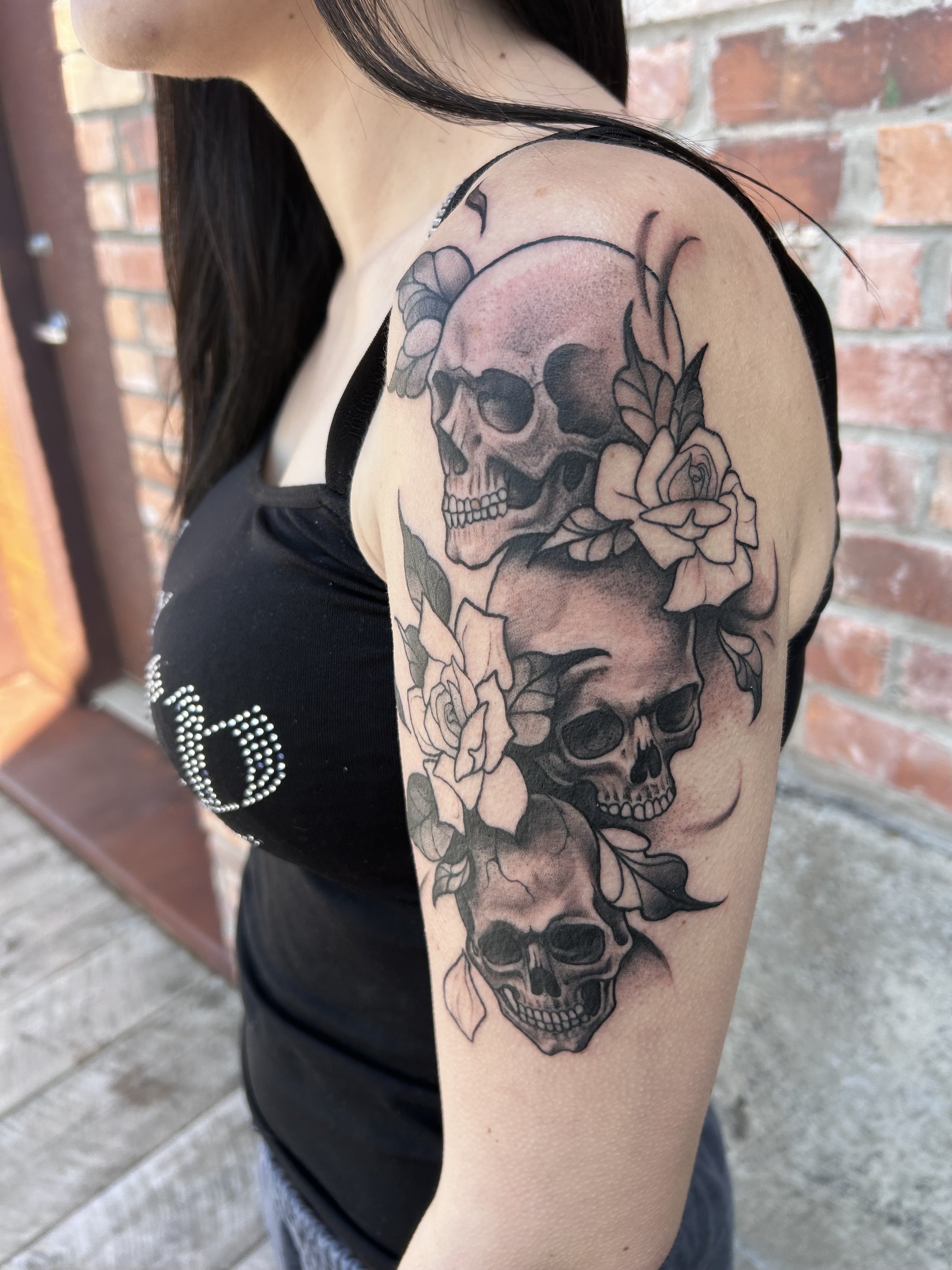 Tattoo of a skull with roses on a person's upper arm, against a brick wall background.