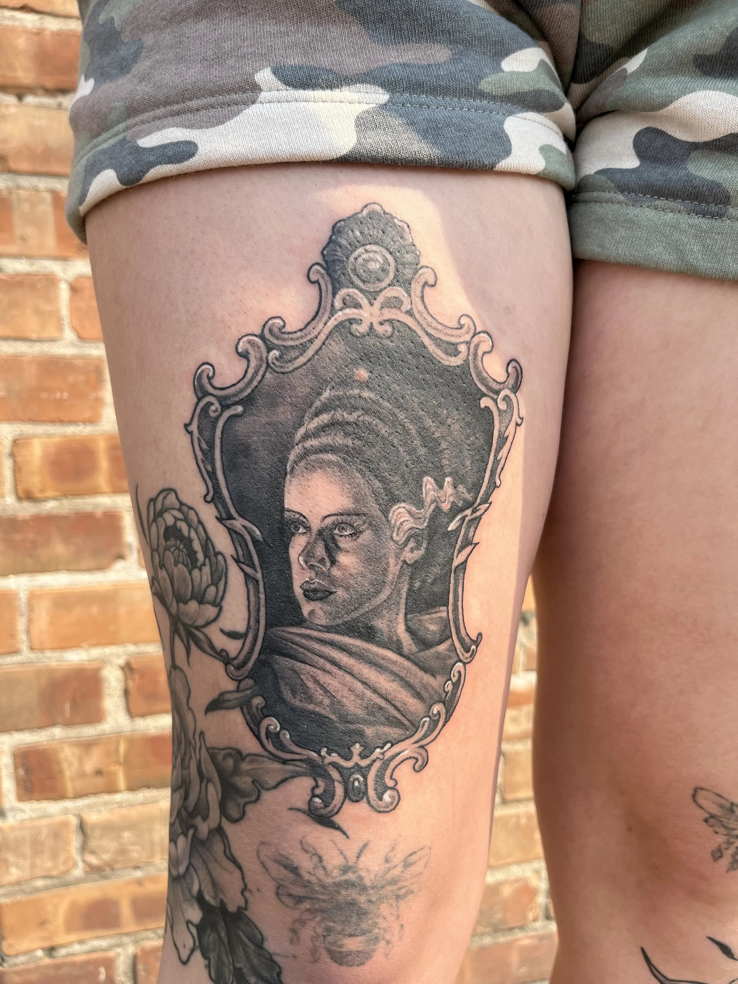 A tattoo of a woman's face inside an ornate decorative frame on a person's thigh, with a brick wall in the background.