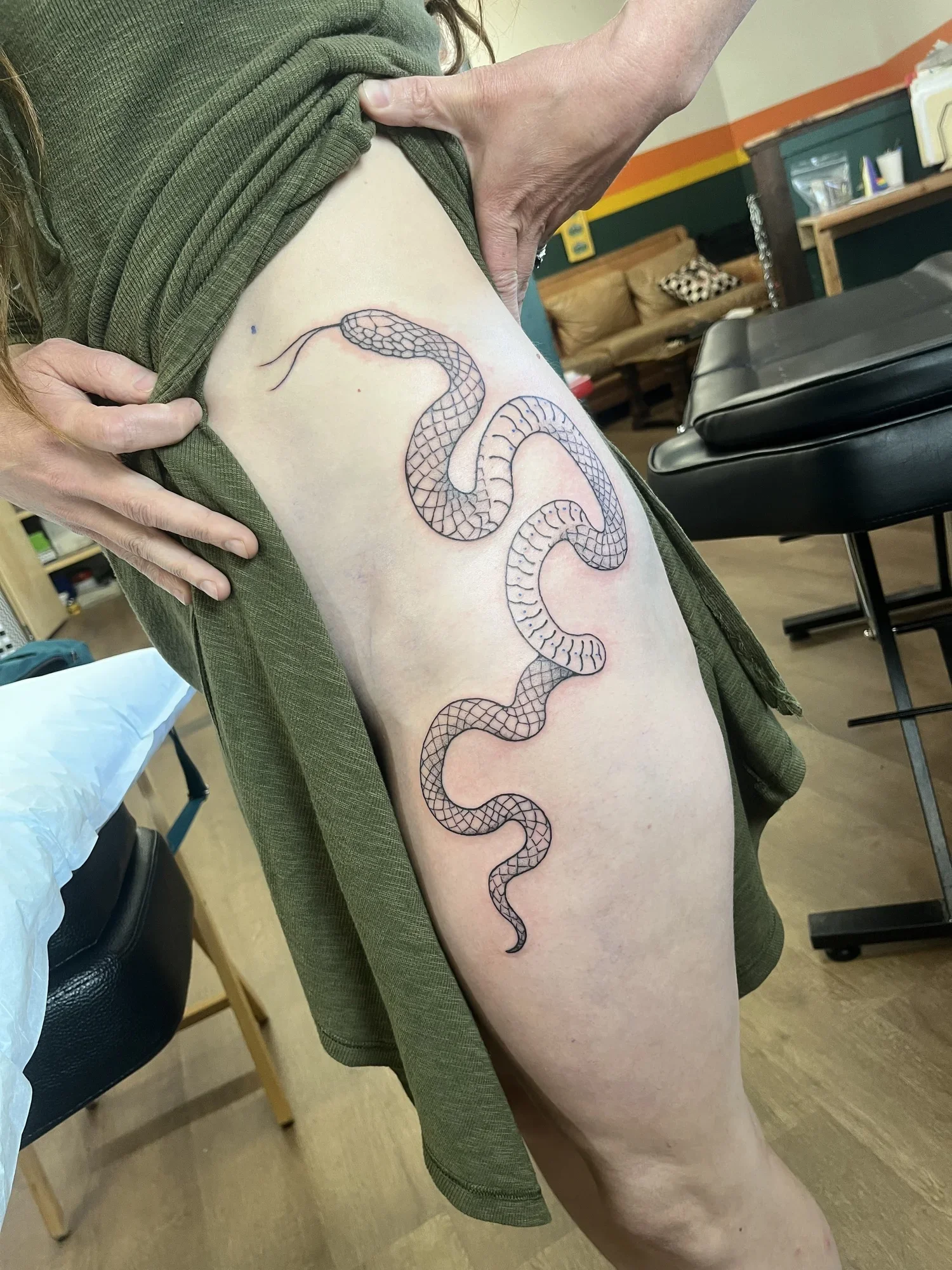 A person showing a fresh snake tattoo on their upper thigh, with their green shirt lifted to reveal it, in a room with chairs, tables, and a couch.
