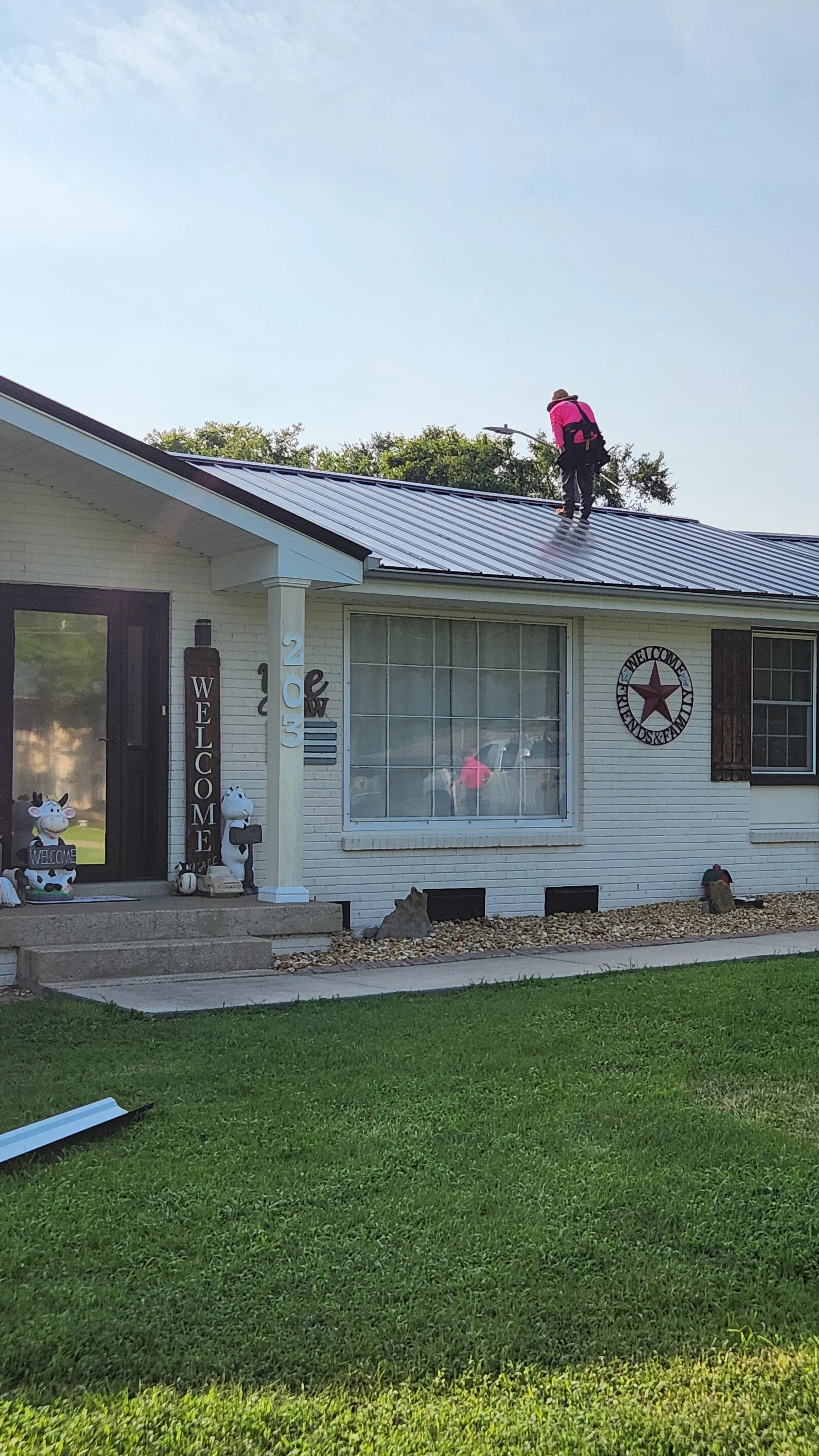 Person wearing pink jacket and hat cleaning a metal roof on a white house with decorations including cow figurines, welcome signs, and American flag-themed items.