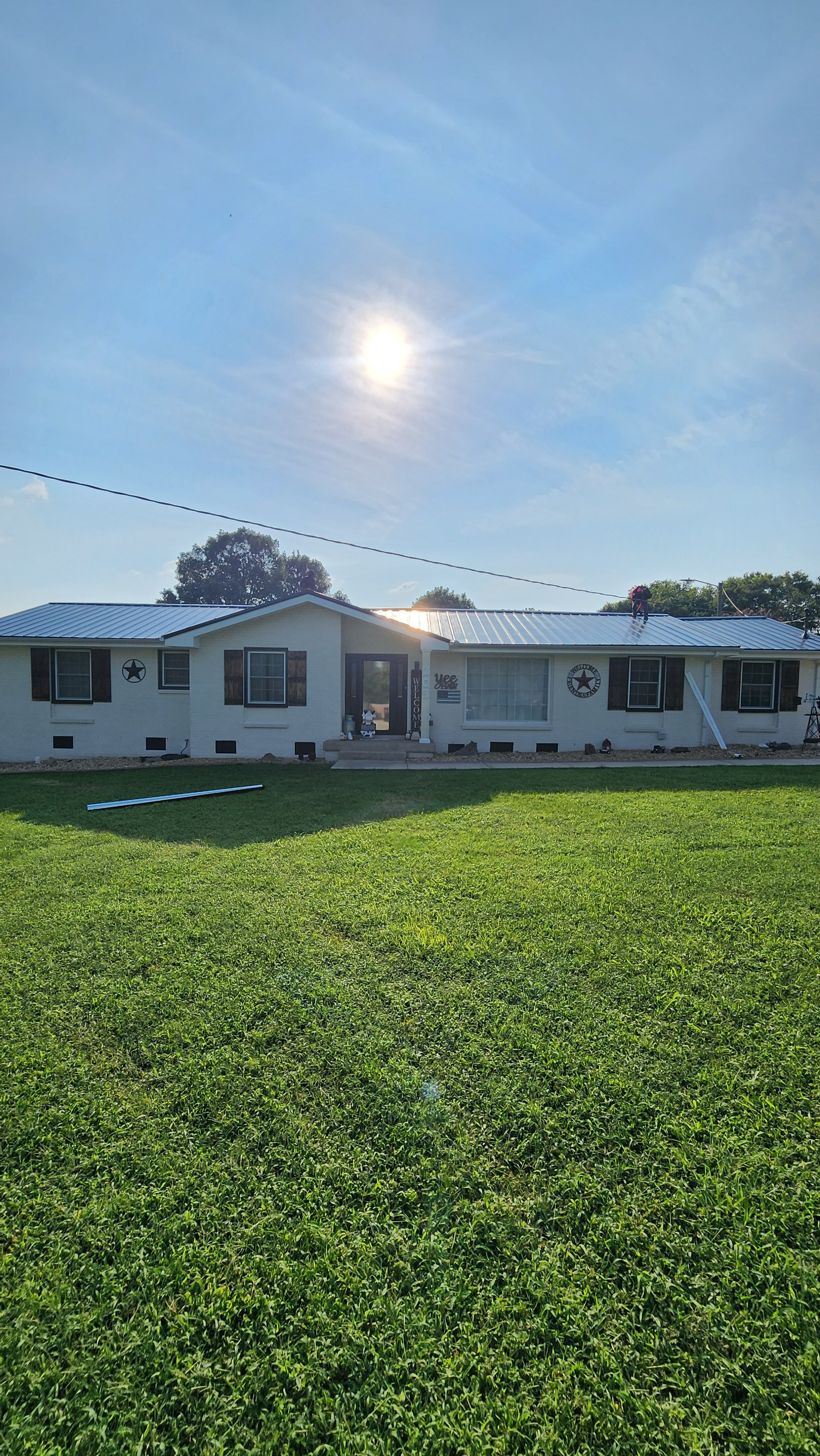 A white house with a metal roof, decorated with star-themed accents, situated in a green lawn under a bright sun in a partly cloudy sky. A person is working on the roof.